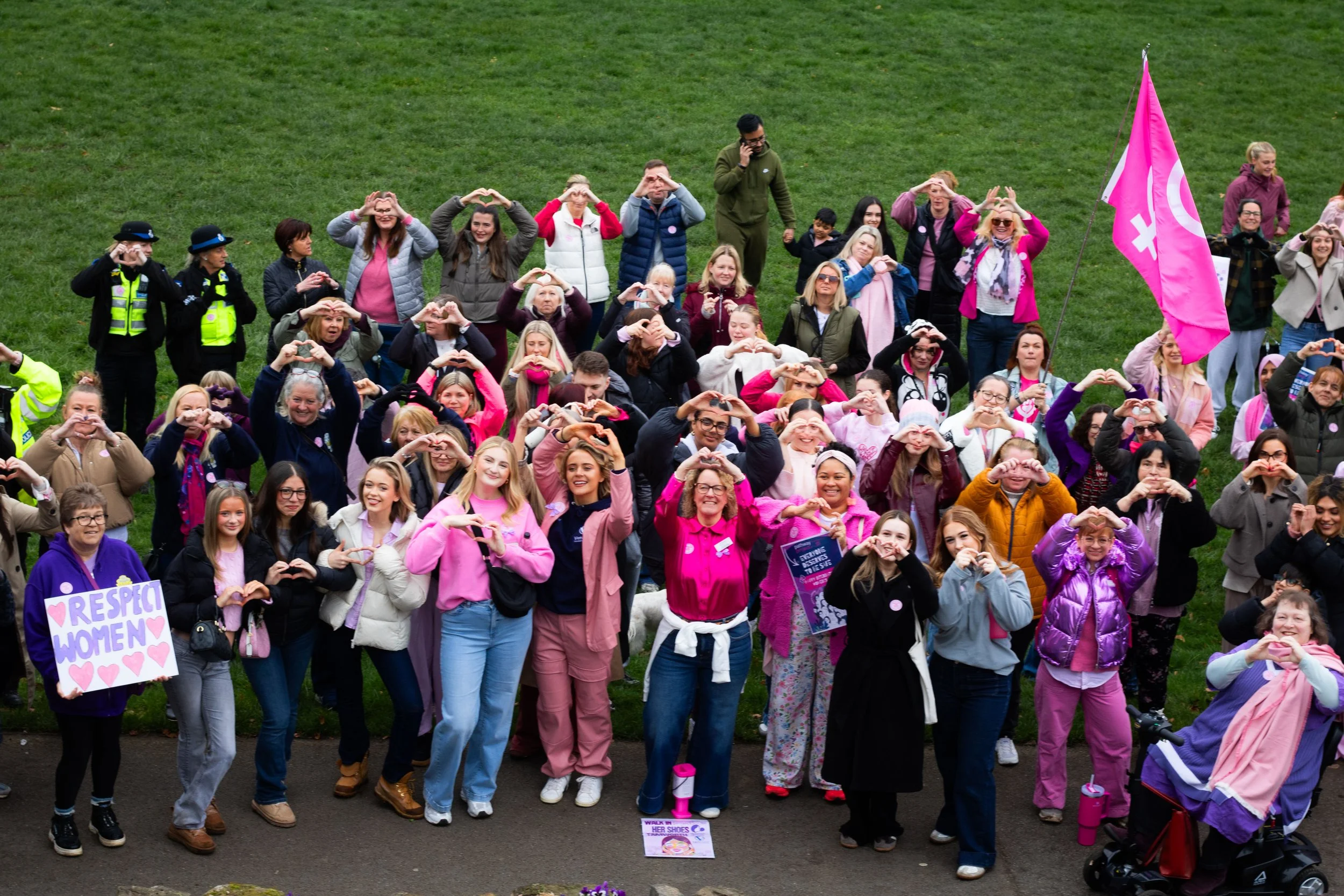 A large group of women and some men gathered on a grassy area, many making heart shapes with their hands. Some participants are wearing pink or purple, and a pink flag is visible on the right side. There are police officers present, and some individuals are holding signs with messages supporting women and respect.