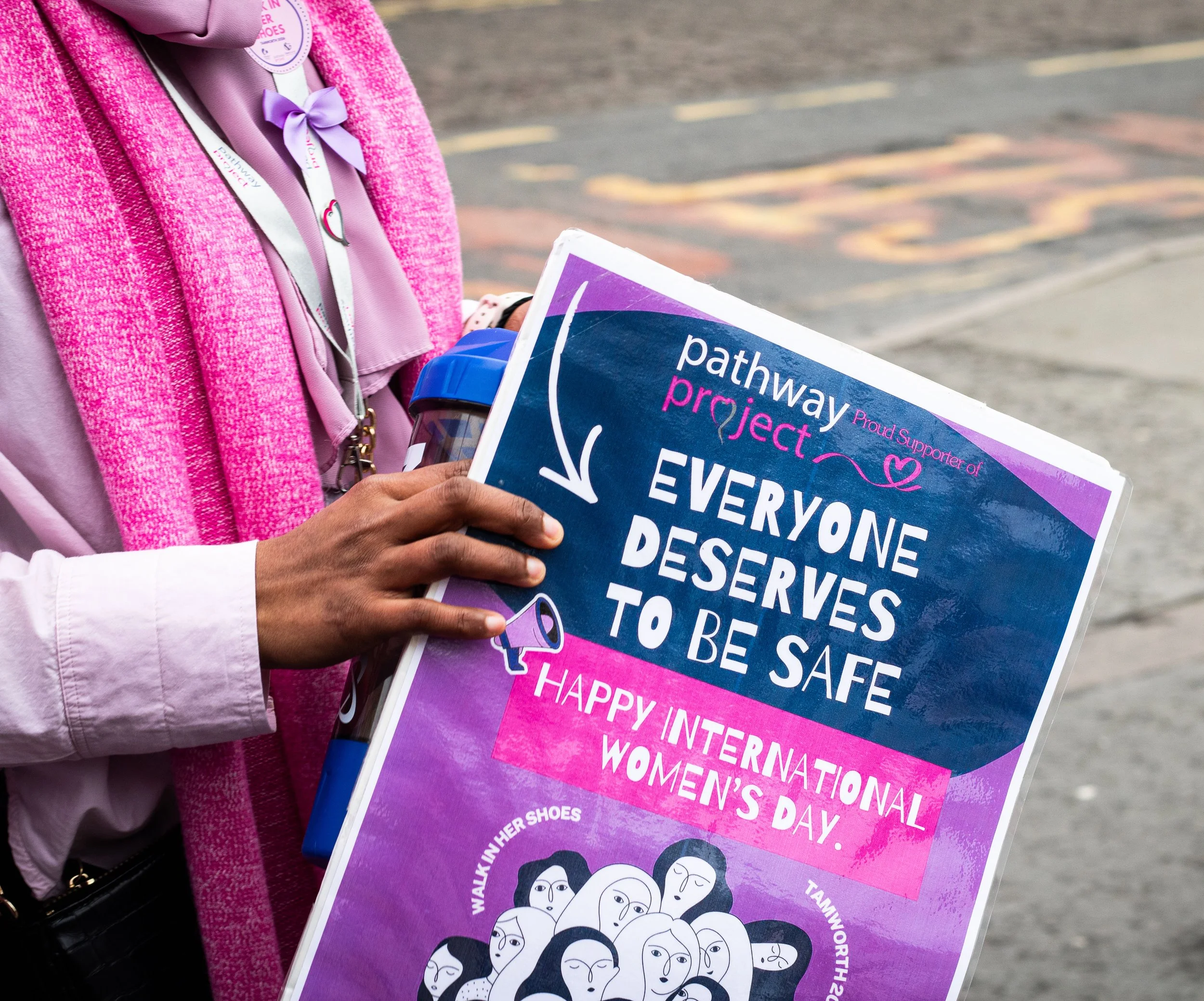 Person holding a sign supporting International Women's Day with pink and purple colors, illustrations of diverse women's faces, and text messages promoting women's rights.