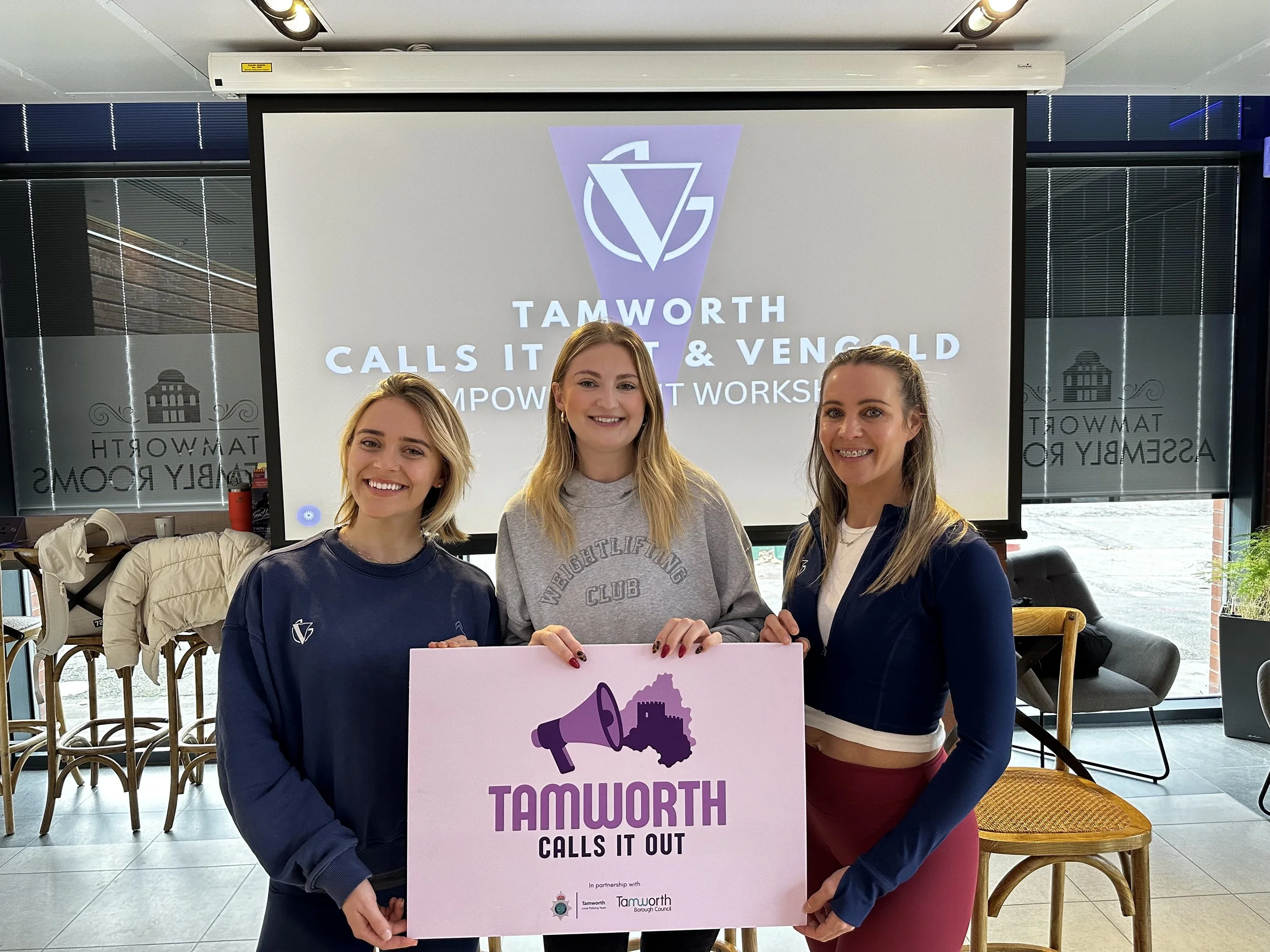 Three women holding a sign that says 'Tamworth Calls It Out' with a megaphone graphic, standing in an indoor space with glass walls, chairs, and a large screen behind them displaying the same message.