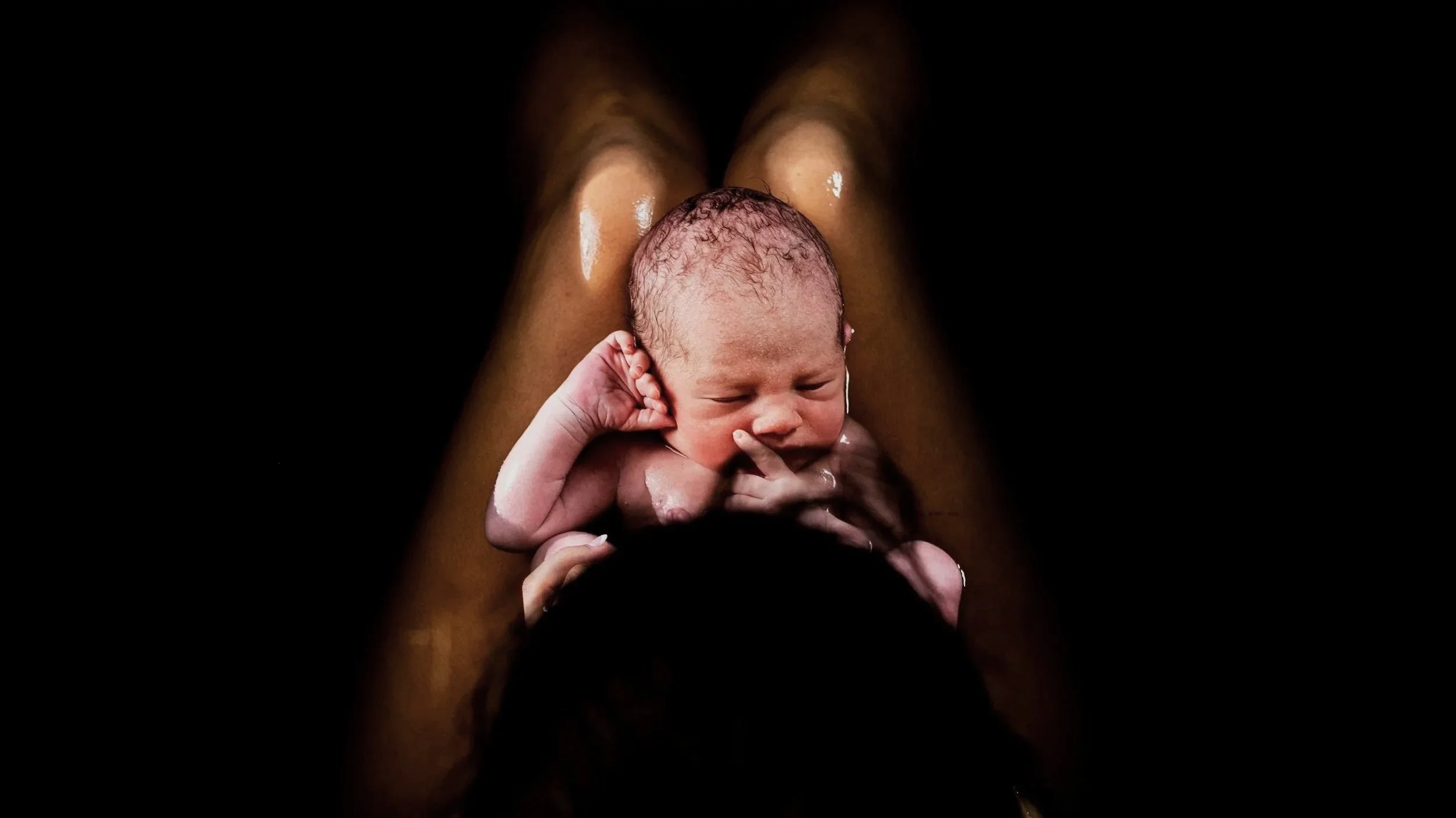 A newborn baby being bathed, held by an adult's hands, with water and skin visible, against a dark background.