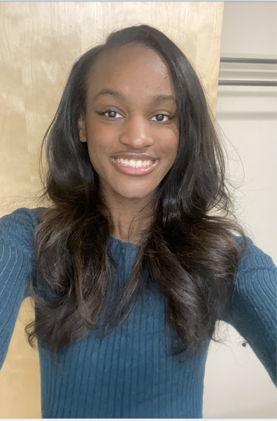 A young woman with dark wavy hair taking a selfie, smiling, wearing a blue ribbed long sleeve shirt, standing in front of a light-colored wooden door and indoor setting.