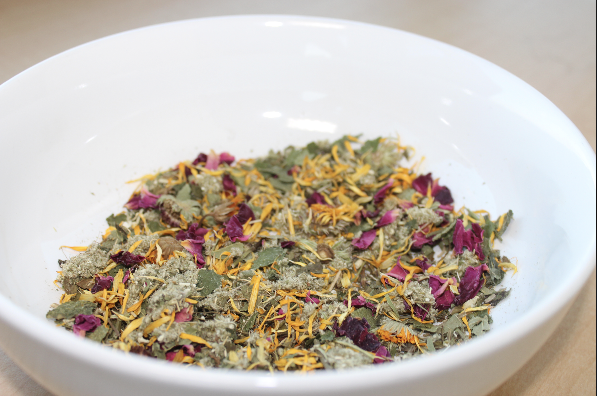 A white bowl containing a mixture of dried herbs and flower petals, including green leaves, purple petals, and yellow, orange petals.