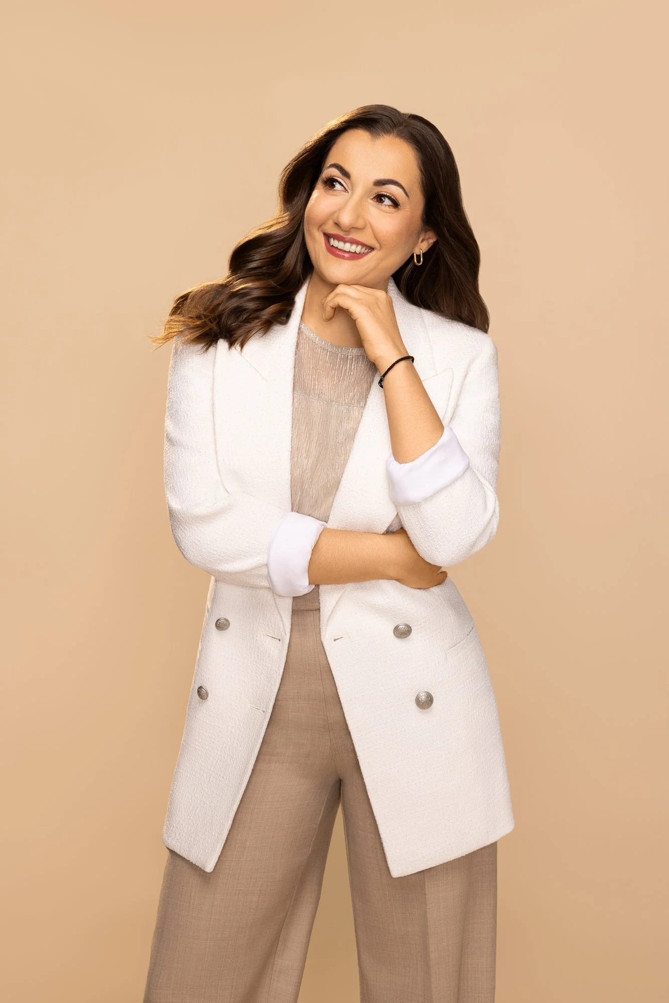 A woman with dark brown wavy hair is smiling and looking to her left, touching her chin with her right hand, against a beige background. She is dressed in a white textured coat over a beige top and matching beige pants, with gold hoop earrings and a black bracelet.