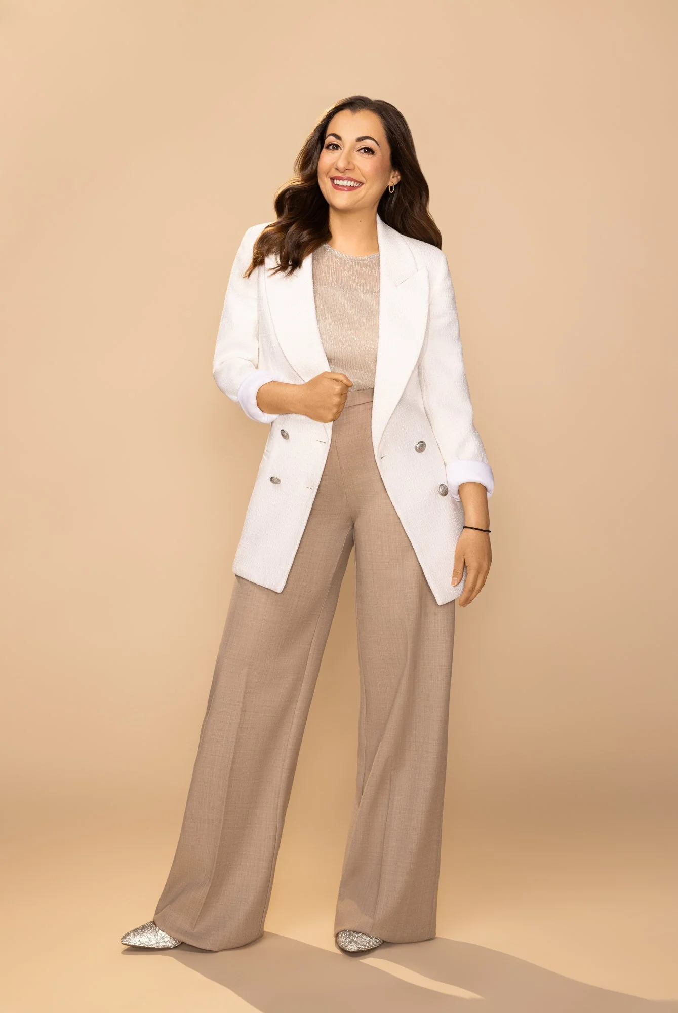 A woman with long brunette hair, wearing a white blazer over a beige top, beige wide-leg pants, and glittery silver shoes, standing against a plain beige background and smiling confidently.