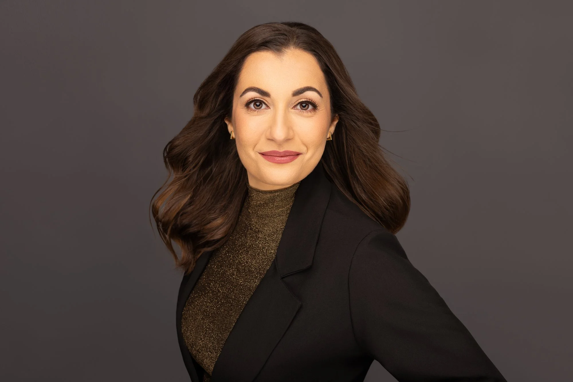 A woman with long wavy brown hair, wearing a black blazer over a glittery gold top, smiling against a plain grey background.