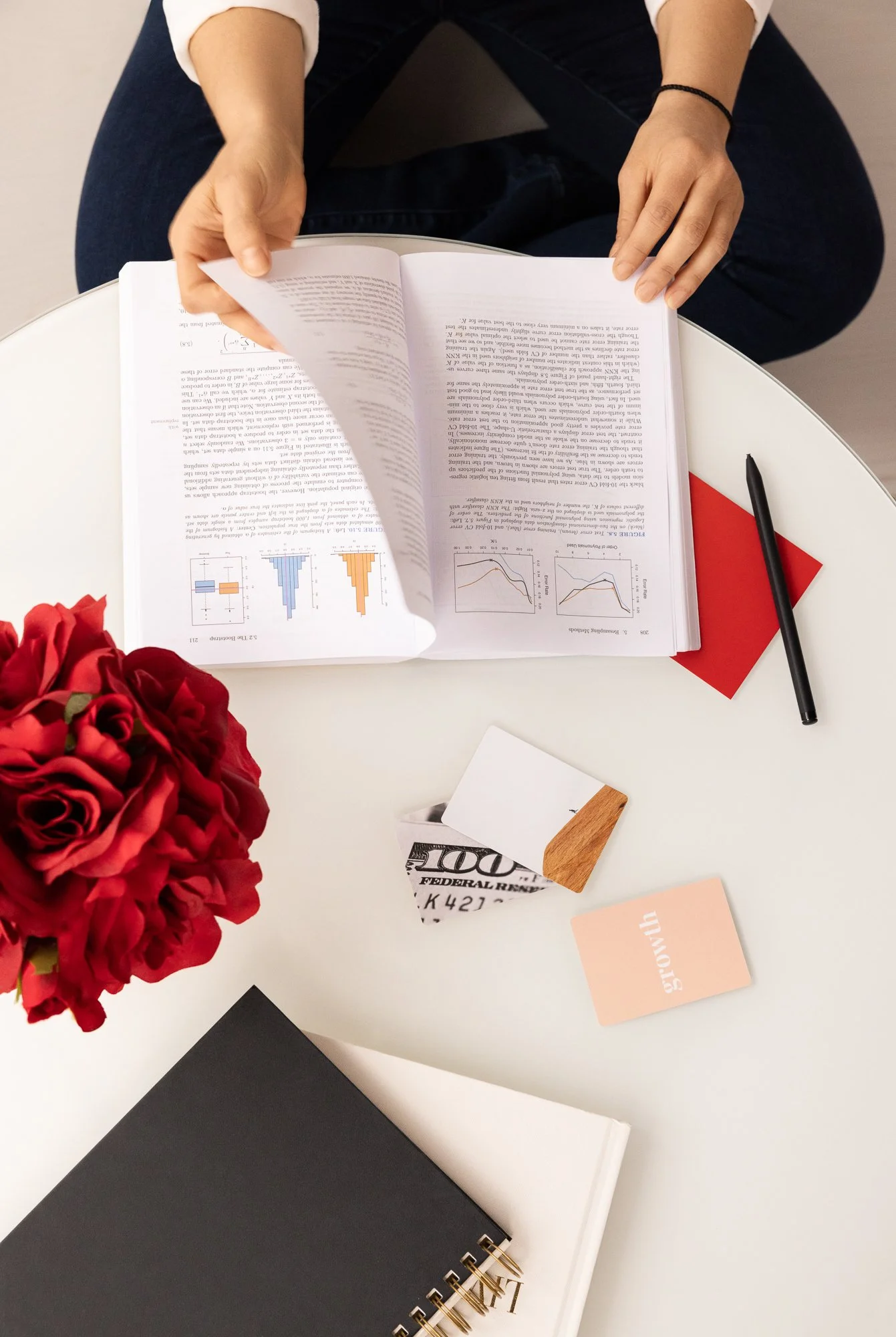 A person sitting at a white table, flipping through a booklet with graphs and text, surrounded by flowers, notebooks, a pen, and small cards on the table.
