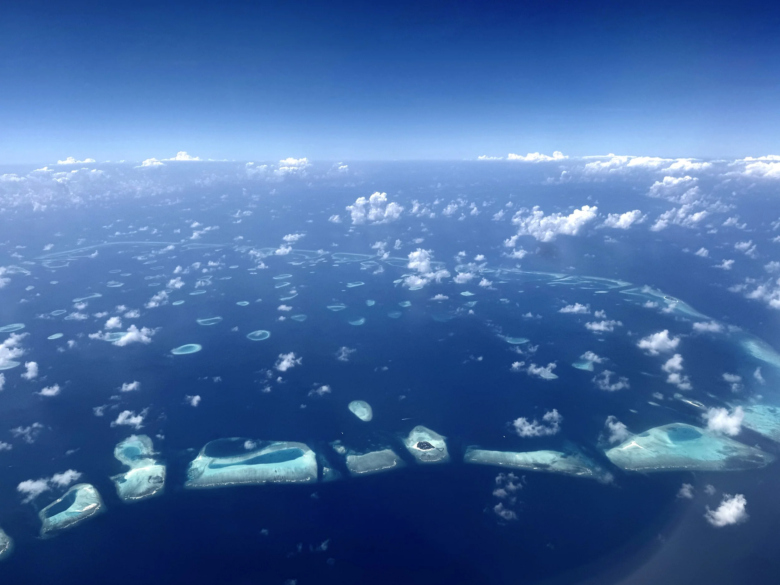 Aerial view of a deep blue ocean with numerous small, circular coral formations visible beneath the water's surface and scattered fluffy white clouds above.