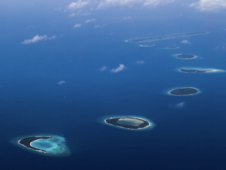 Aerial view of multiple coral atolls in the ocean with clear blue water and clouds above.