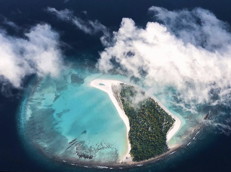 Aerial view of a tropical island with a white sandy beach, lush green trees, and surrounding turquoise waters, partially obscured by clouds.