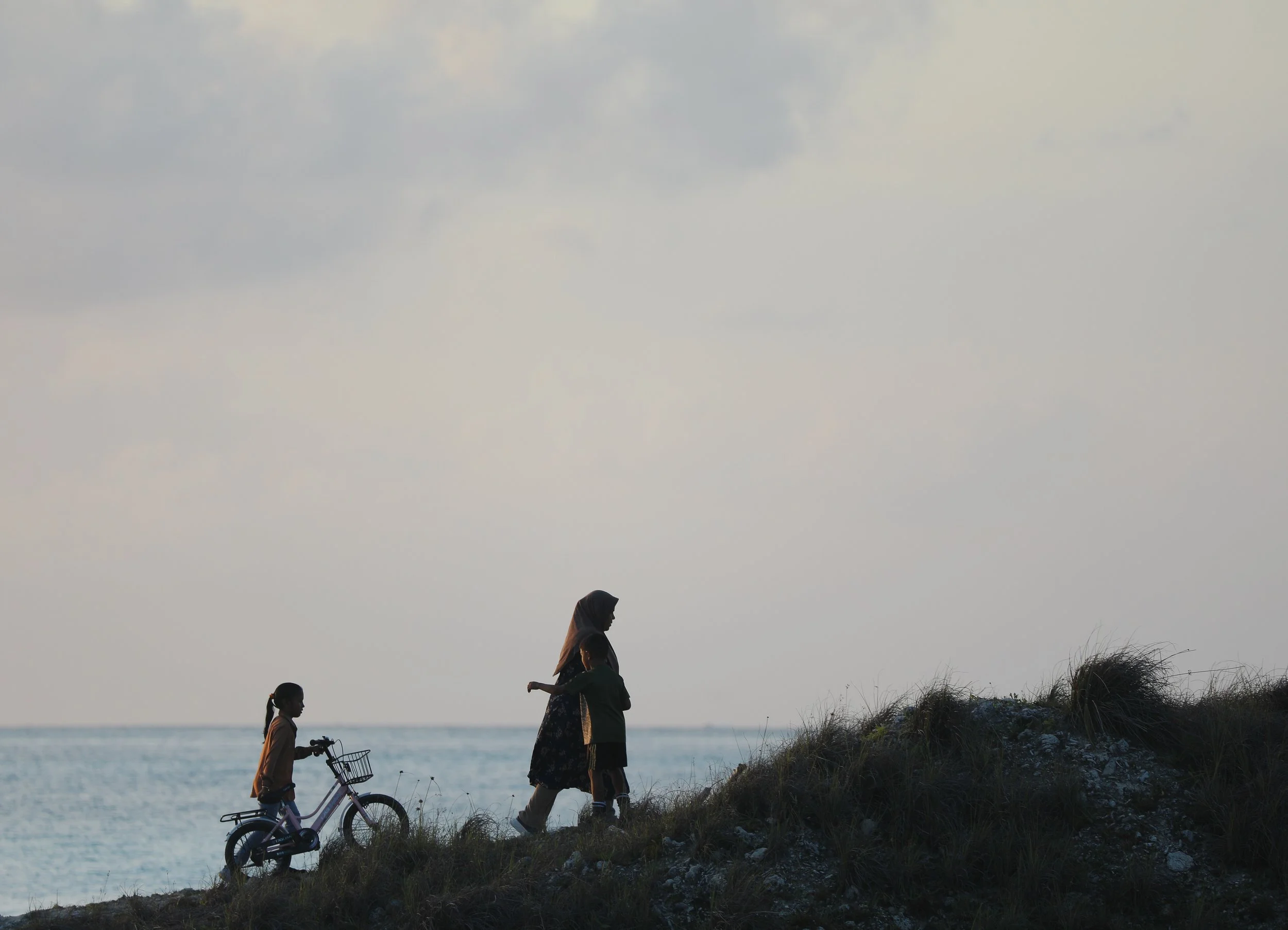 A woman and a girl walking along a hill by the water, with the girl riding a bicycle. The woman is wearing a headscarf, and the girl has pigtails. The sky is slightly clouded.