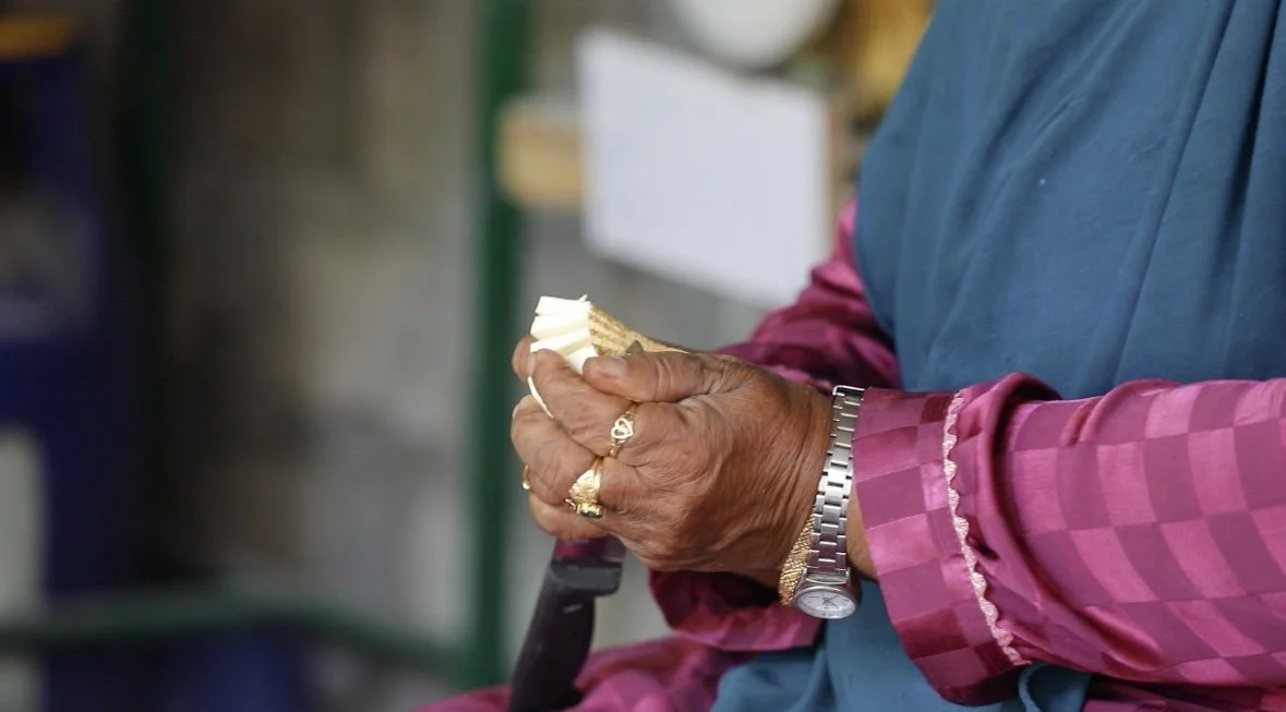 Close-up of an elderly person's hands holding a partially peeled potato, wearing jewelry and a blue and pink checkered shirt.
