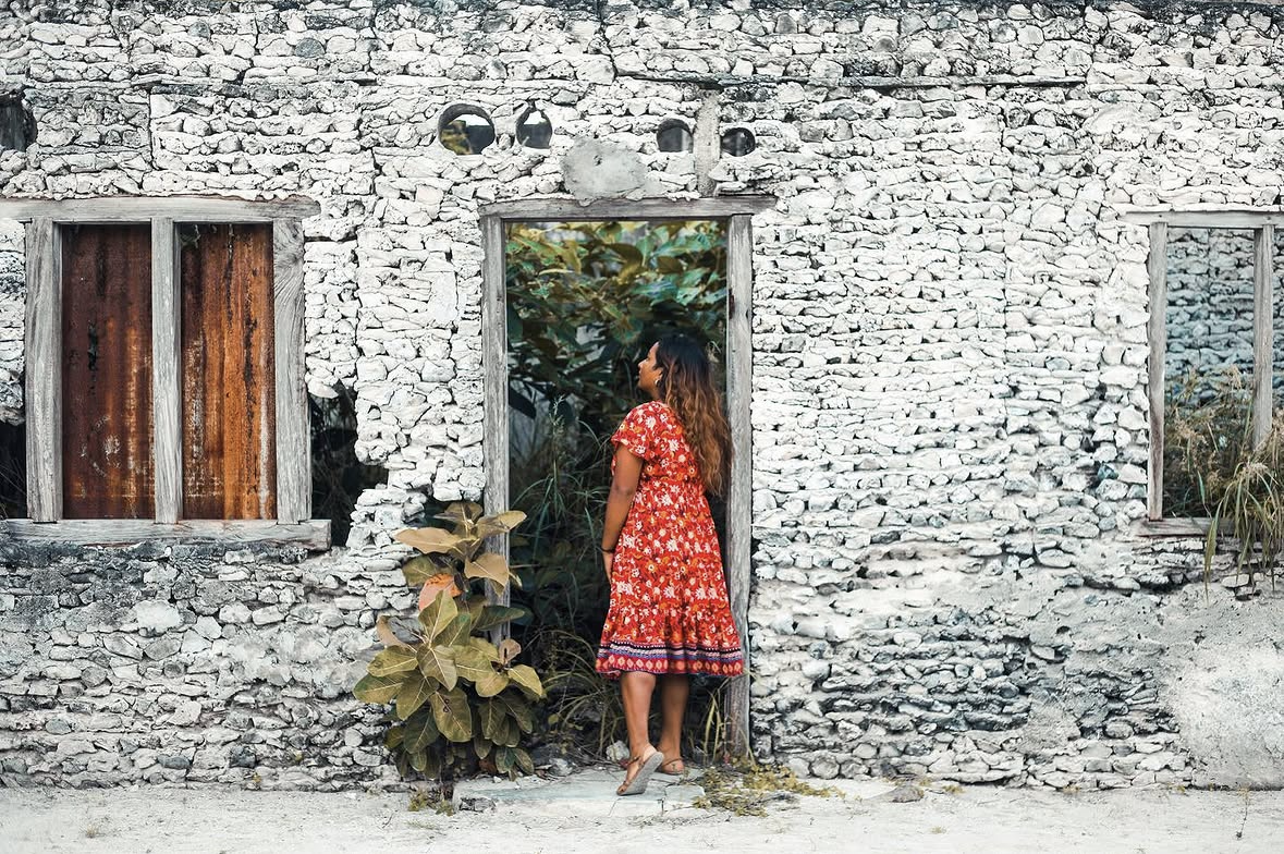 Woman in a flowing red dress standing beside a weathered stone building in a sunlit tropical setting.