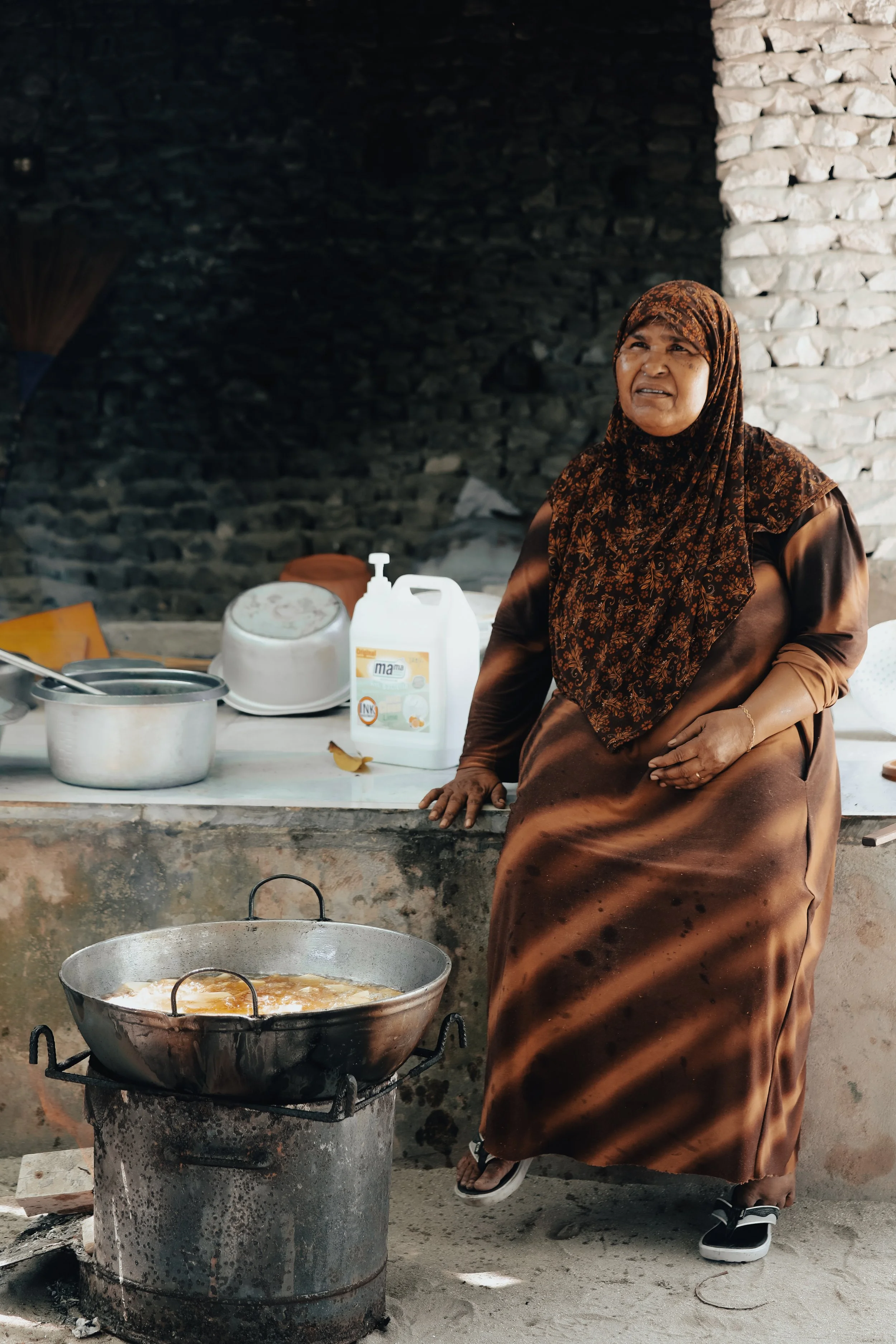 A woman in a brown dress and a patterned headscarf stands beside a large outdoor cooking pot over an open flame, with a brick wall in the background.
