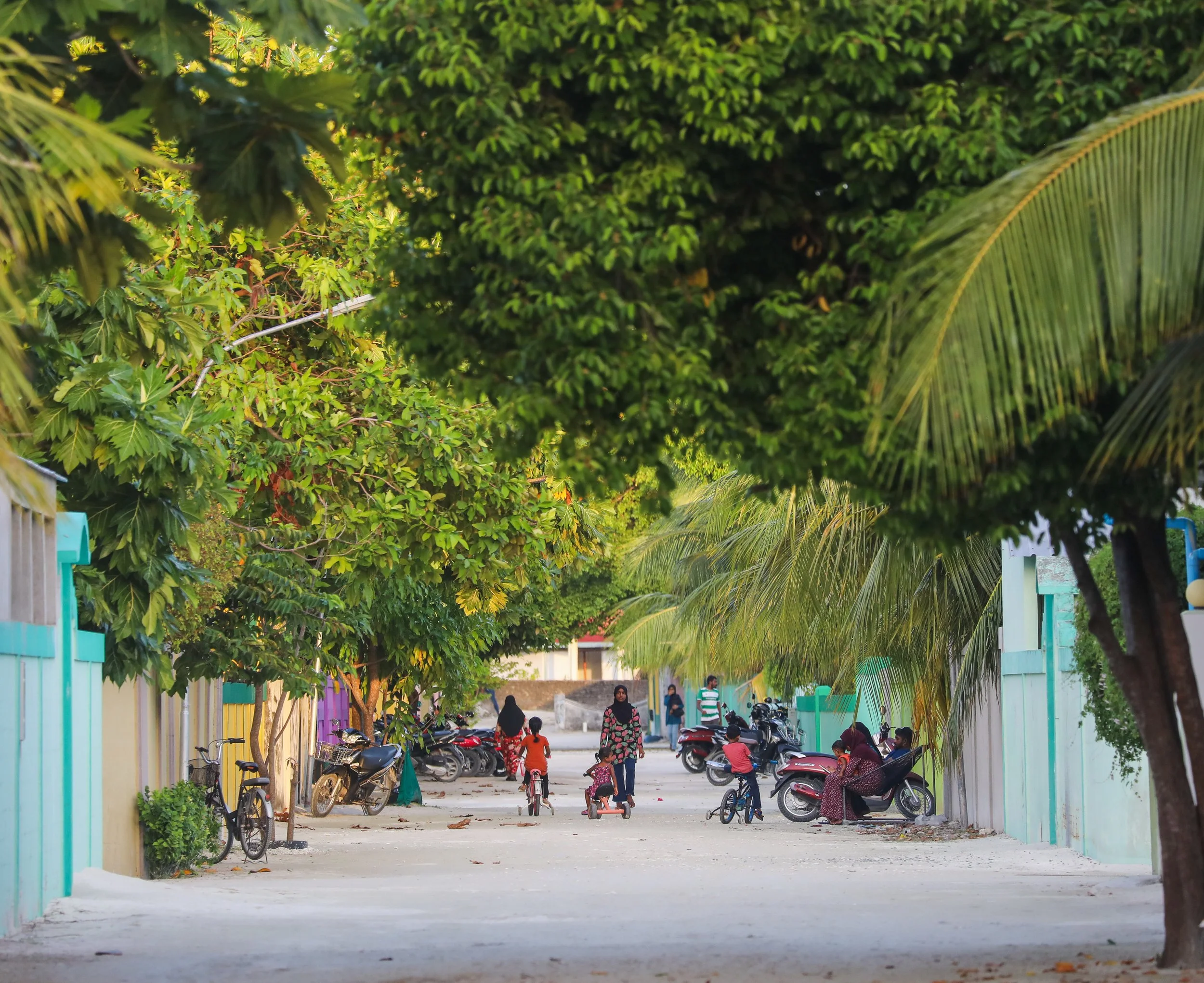 A lively street scene with children riding bikes and playing, adults walking, and parked motorcycles along the colorful buildings, all shaded by green trees.