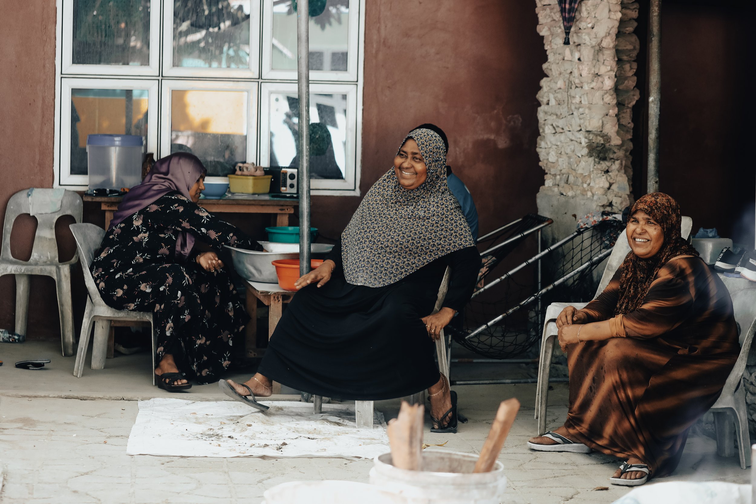 Four women sitting outdoors, laughing and chatting, with a brick wall, window, and kitchen appliances in the background.