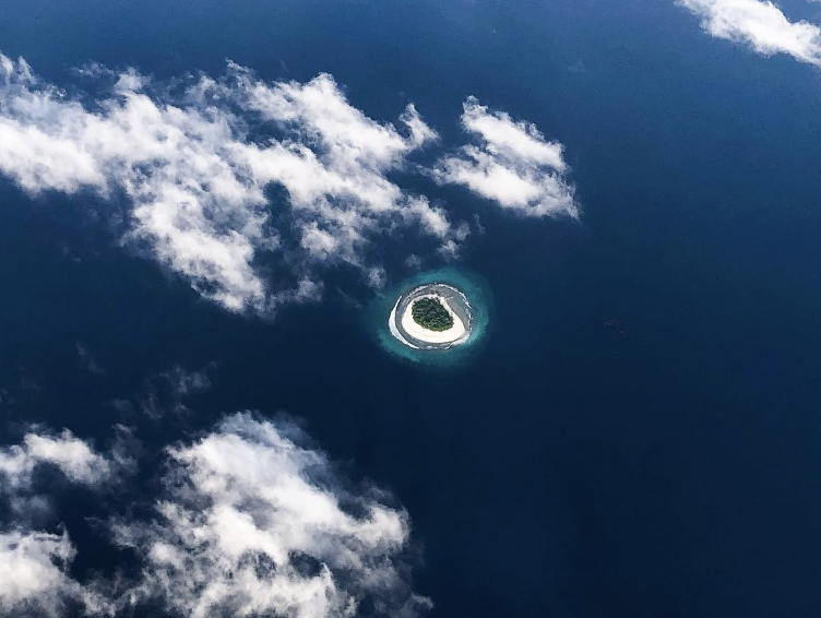Aerial view of a small tropical island with a white sandy beach, surrounded by blue ocean and a few clouds in the sky.