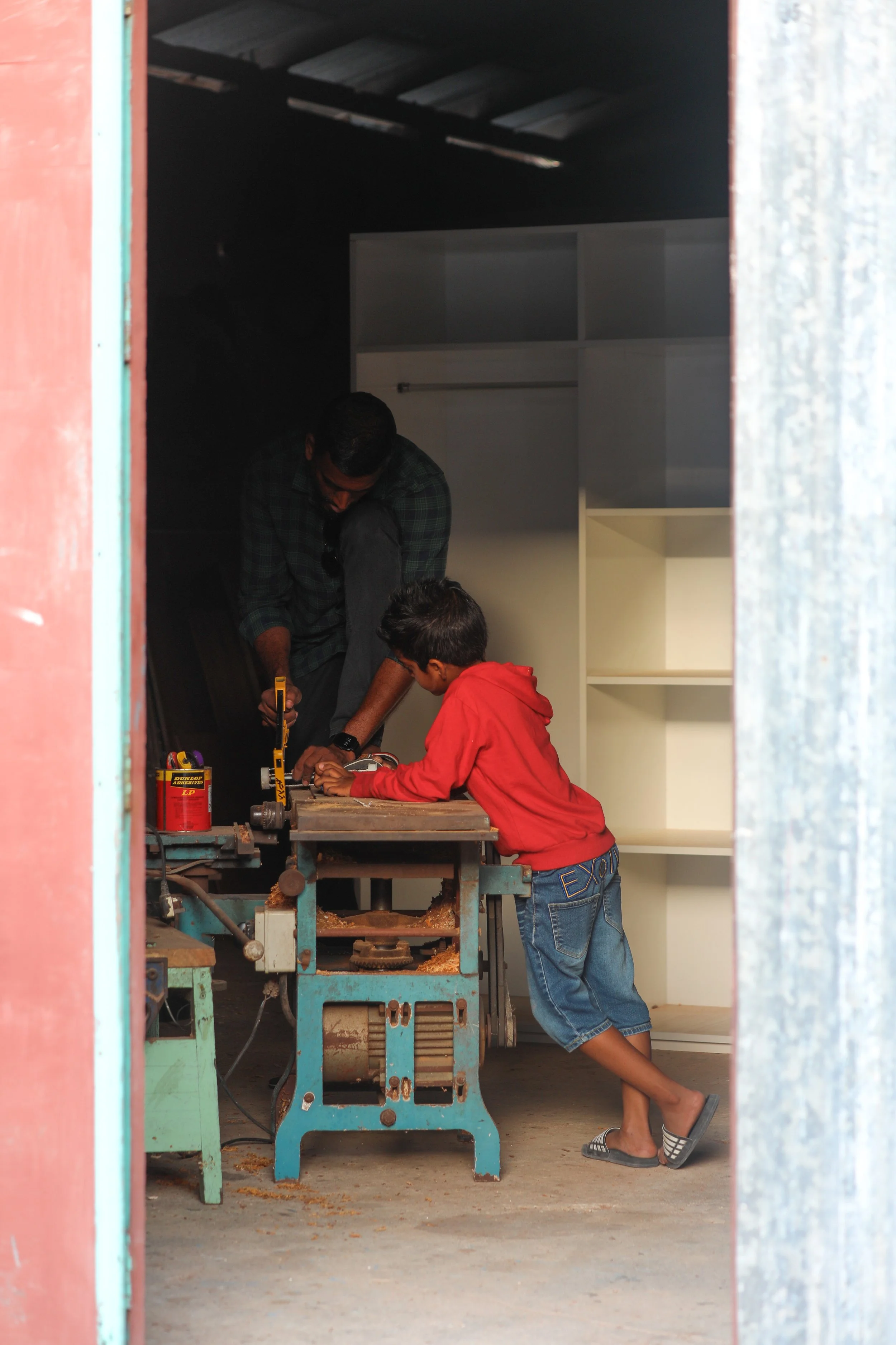 Two children and an adult working together on a woodworking project inside a workshop, viewed through a doorway.