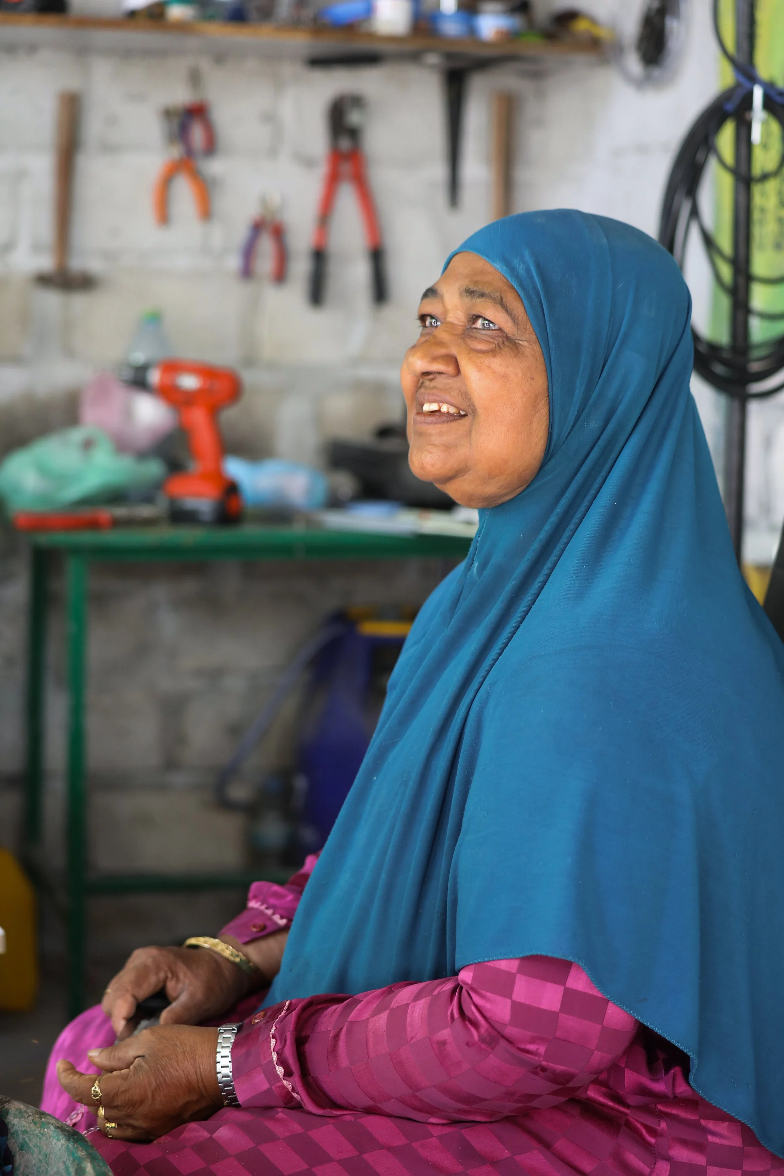 An elderly woman wearing a blue headscarf and a pink checked traditional dress is sitting in a workshop, holding tools in her hands with a cheerful expression.