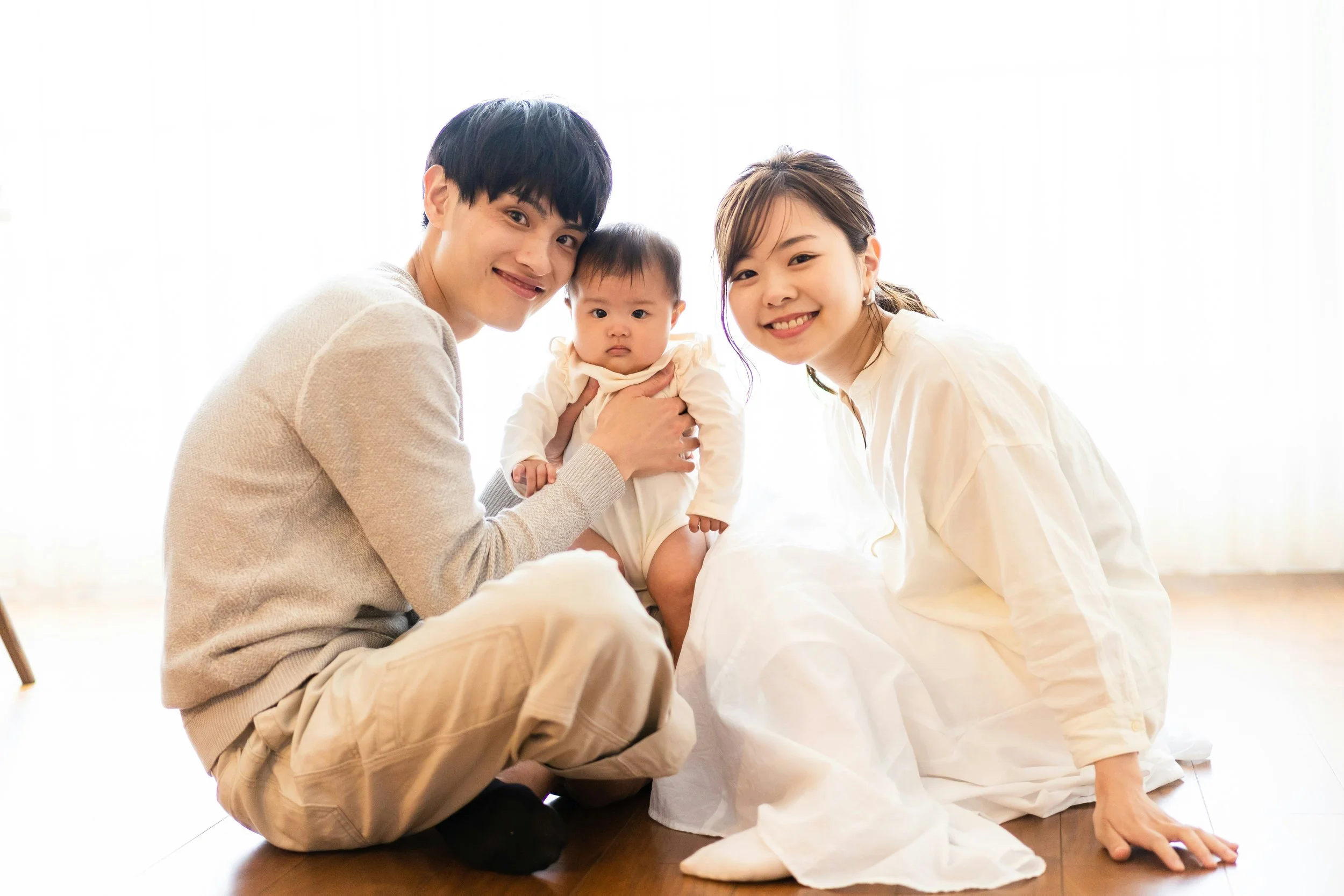 A happy family of three, including a man, a woman, and a baby, sitting together on the floor in bright, natural lighting.