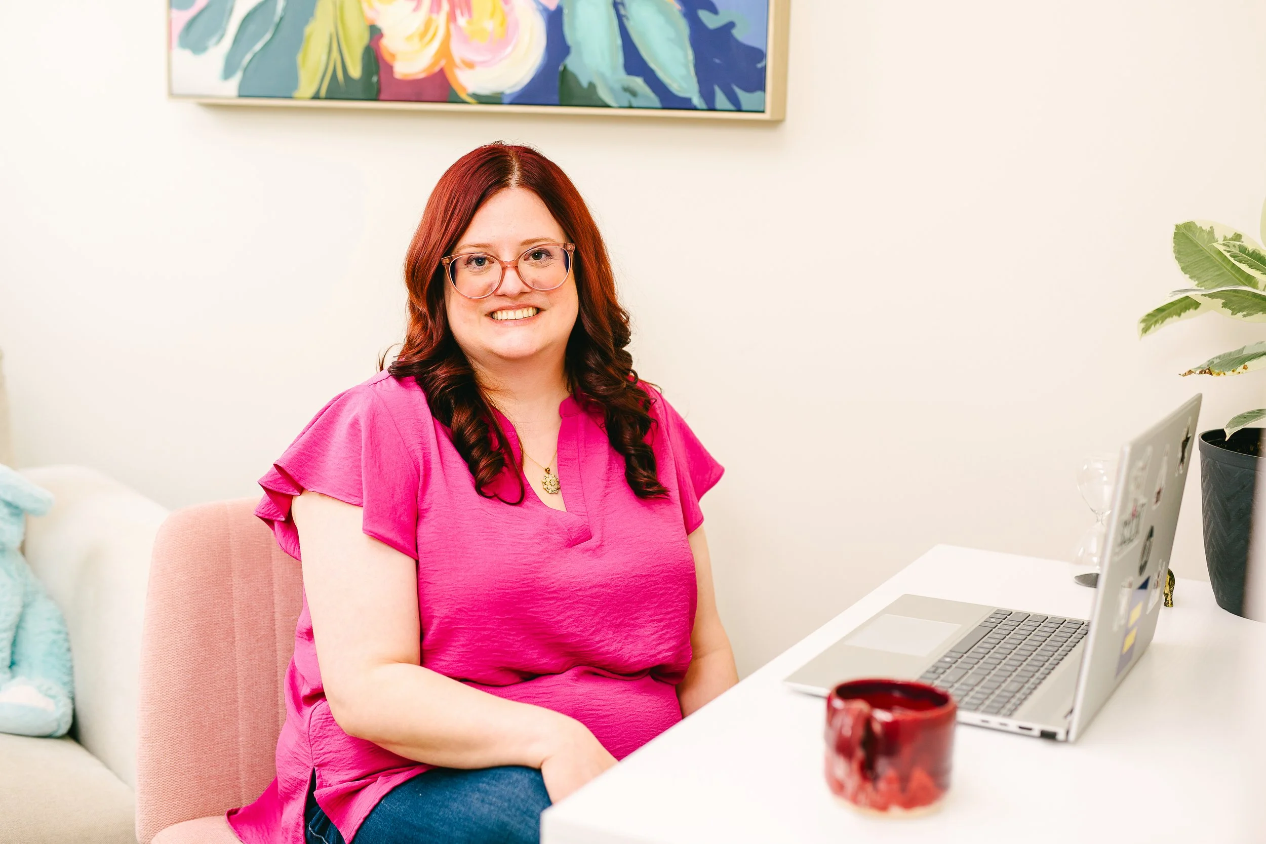 A woman with red hair and glasses smiling while sitting at a white desk with a laptop, a red mug, and some decorative items in a bright room with a white wall, colorful artwork, and a green plant.