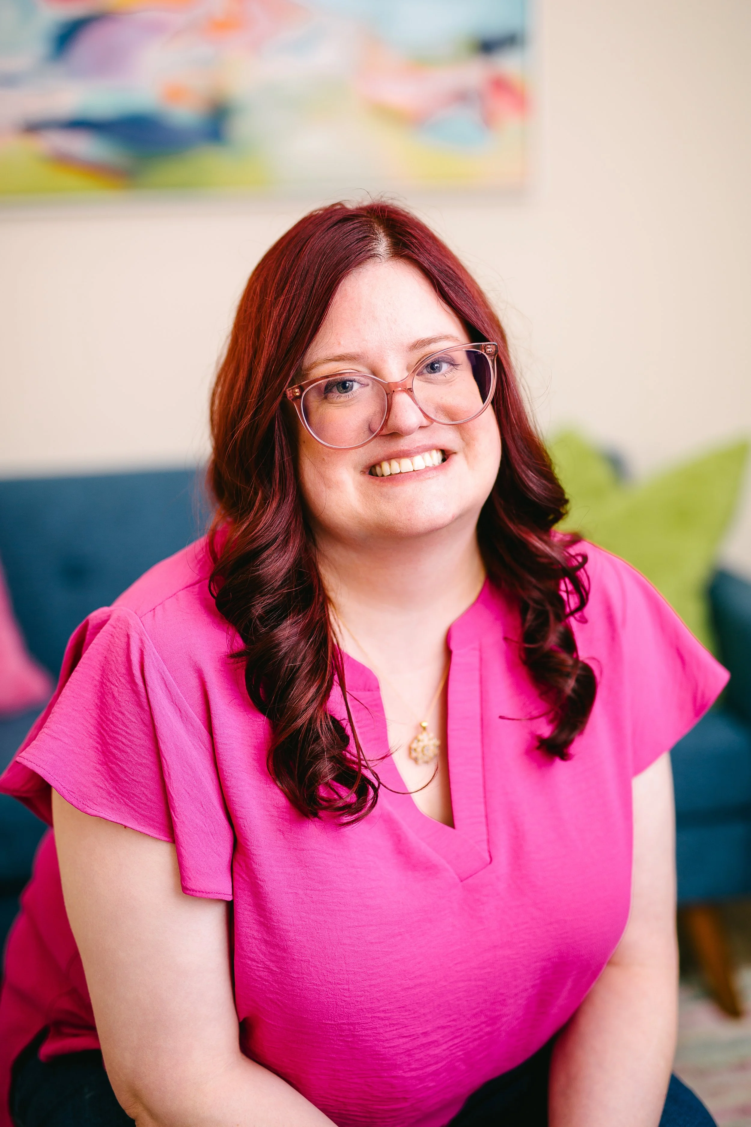 A woman smiling, wearing glasses and a bright pink blouse, sitting indoors with a colorful abstract artwork on the wall behind her.