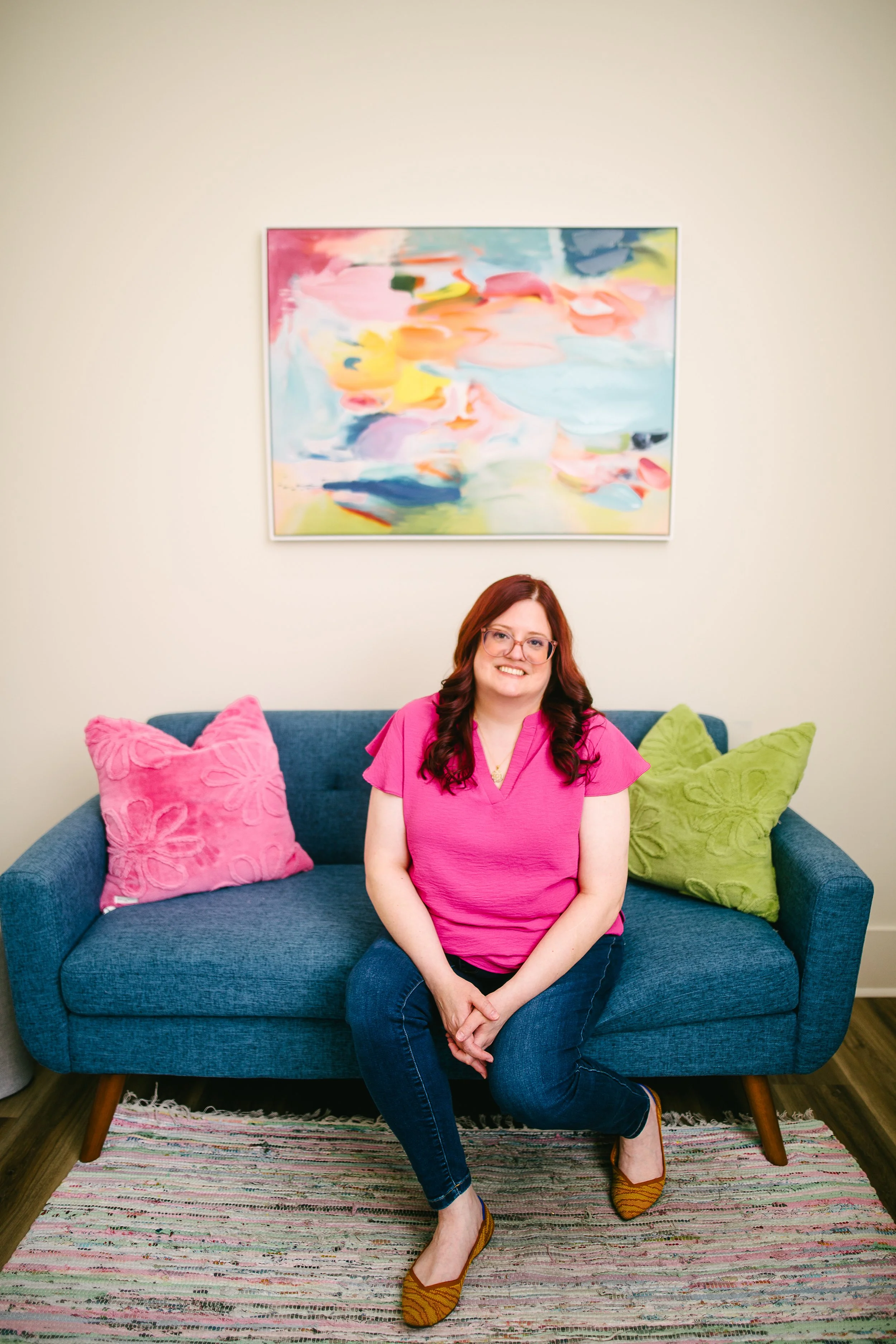 A woman with red hair, glasses, and a pink shirt sitting on a blue couch with pink, green, and black pillows in a living room with a colorful abstract painting on a beige wall and a multi-colored striped rug on the floor.
