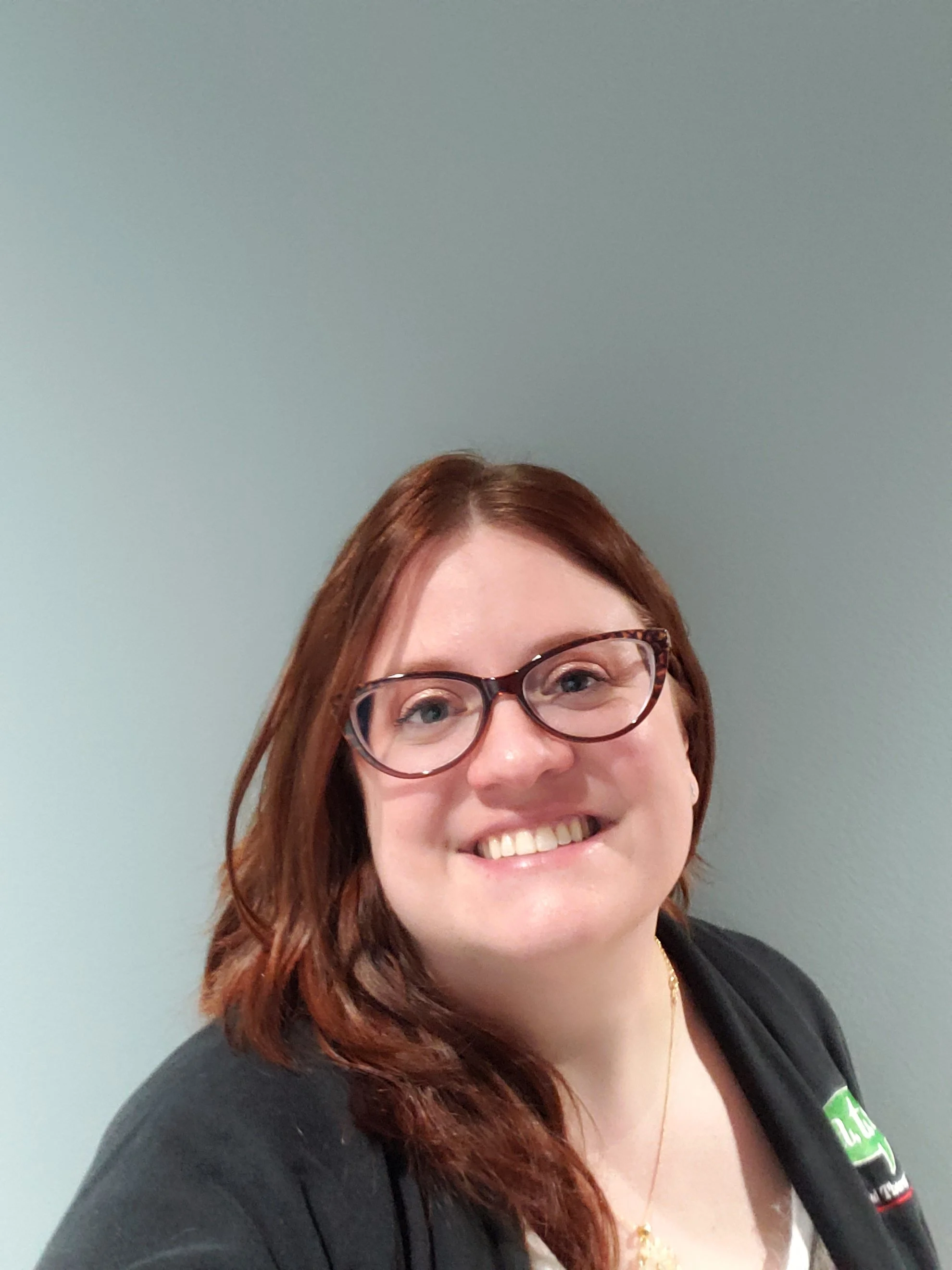 A woman with red hair, wearing glasses, smiling, wearing a black jacket and a white top, standing in front of a plain gray wall.