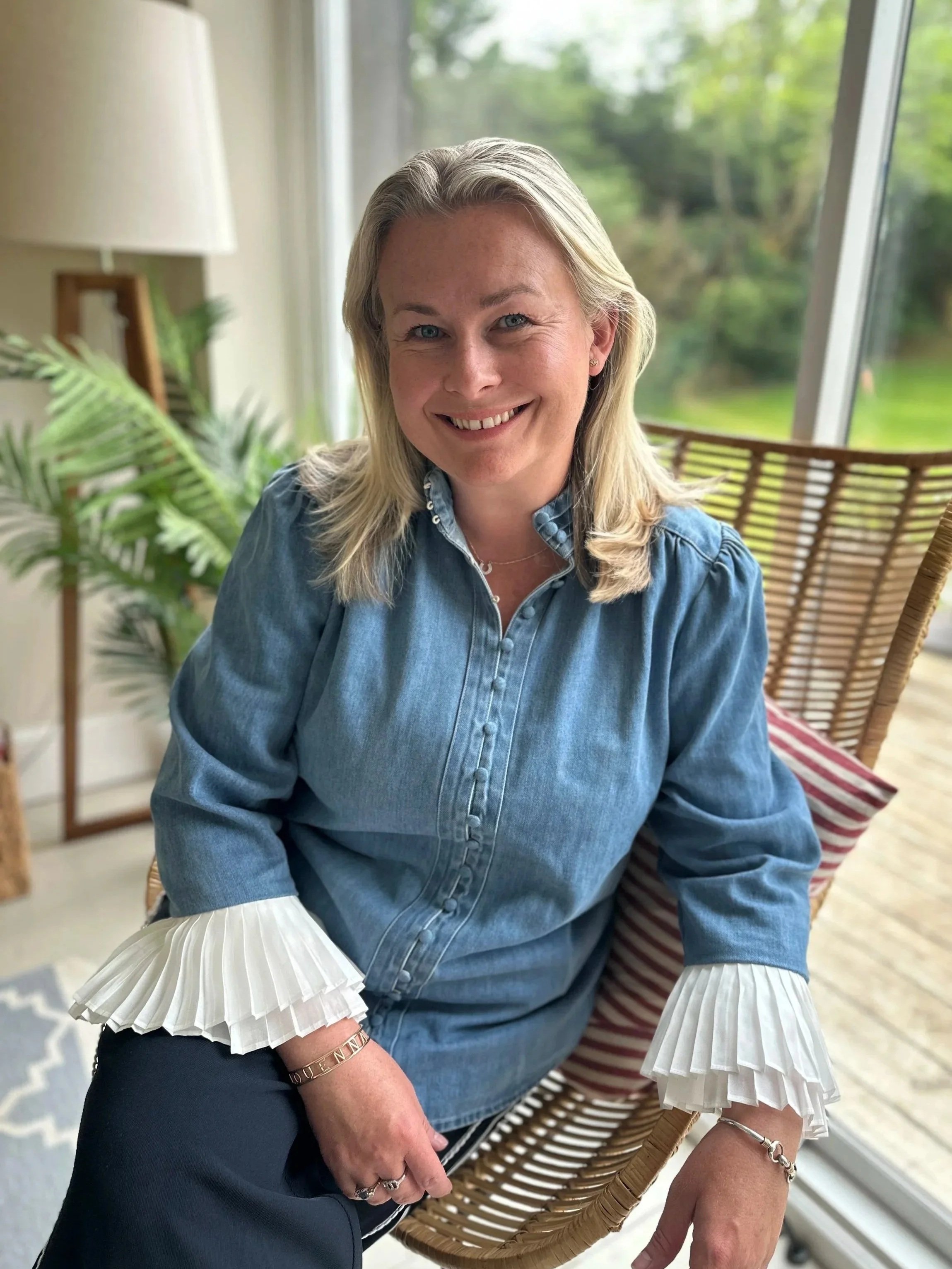 A woman with blonde hair smiling sitting in a wicker chair near a large window with greenery outside, wearing a denim shirt with pleated white sleeves, gold jewelry, and black pants.