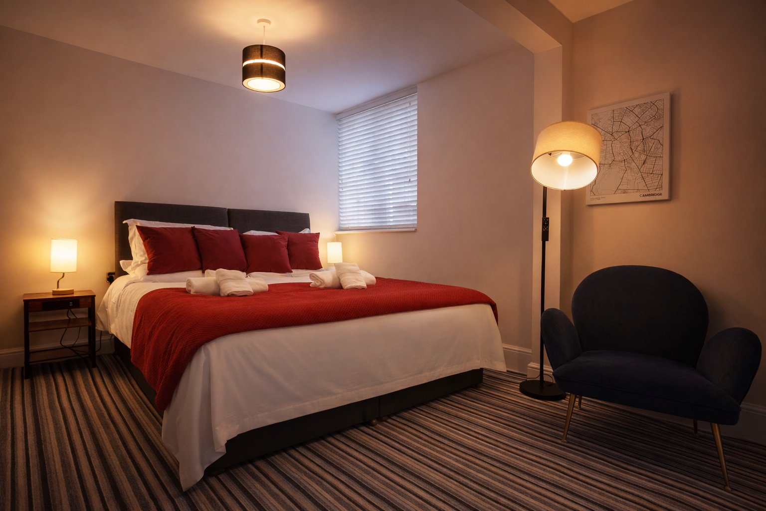 A cozy hotel bedroom with a neatly made bed featuring white sheets, a red bedspread, and multiple red pillows. There are two small lamps on bedside tables, a black armchair with wooden legs, and a tall floor lamp with a white shade. A window with clo