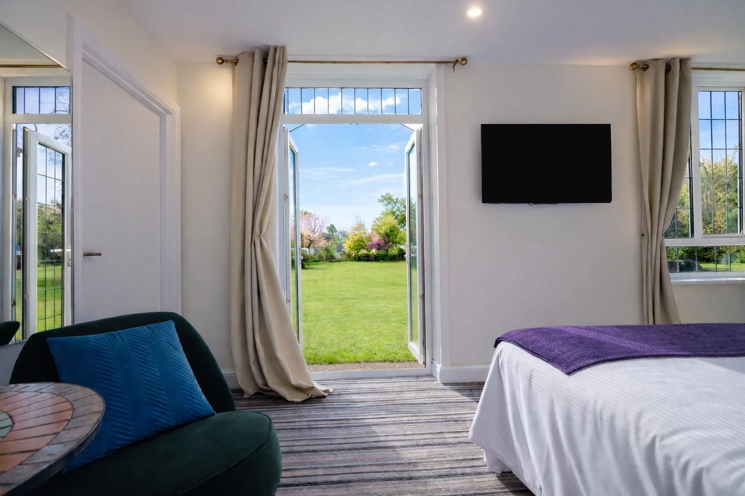 A bright bedroom with open glass doors leading to a green lawn and garden. The room has white walls, beige curtains, a wall-mounted flat-screen TV, a bed with white linens and a purple throw, a dark green chair with a blue pillow, and a partial view 