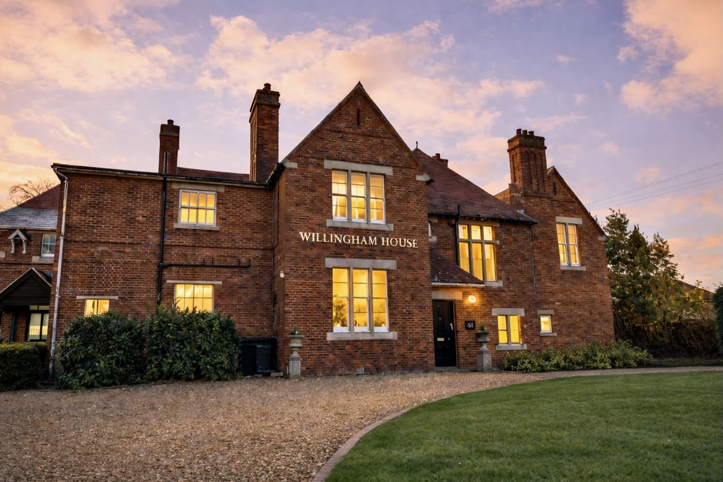 A large brick house with illuminated windows at sunset, labeled 'WILLINGHAM HOUSE,' surrounded by a gravel driveway and green lawn.