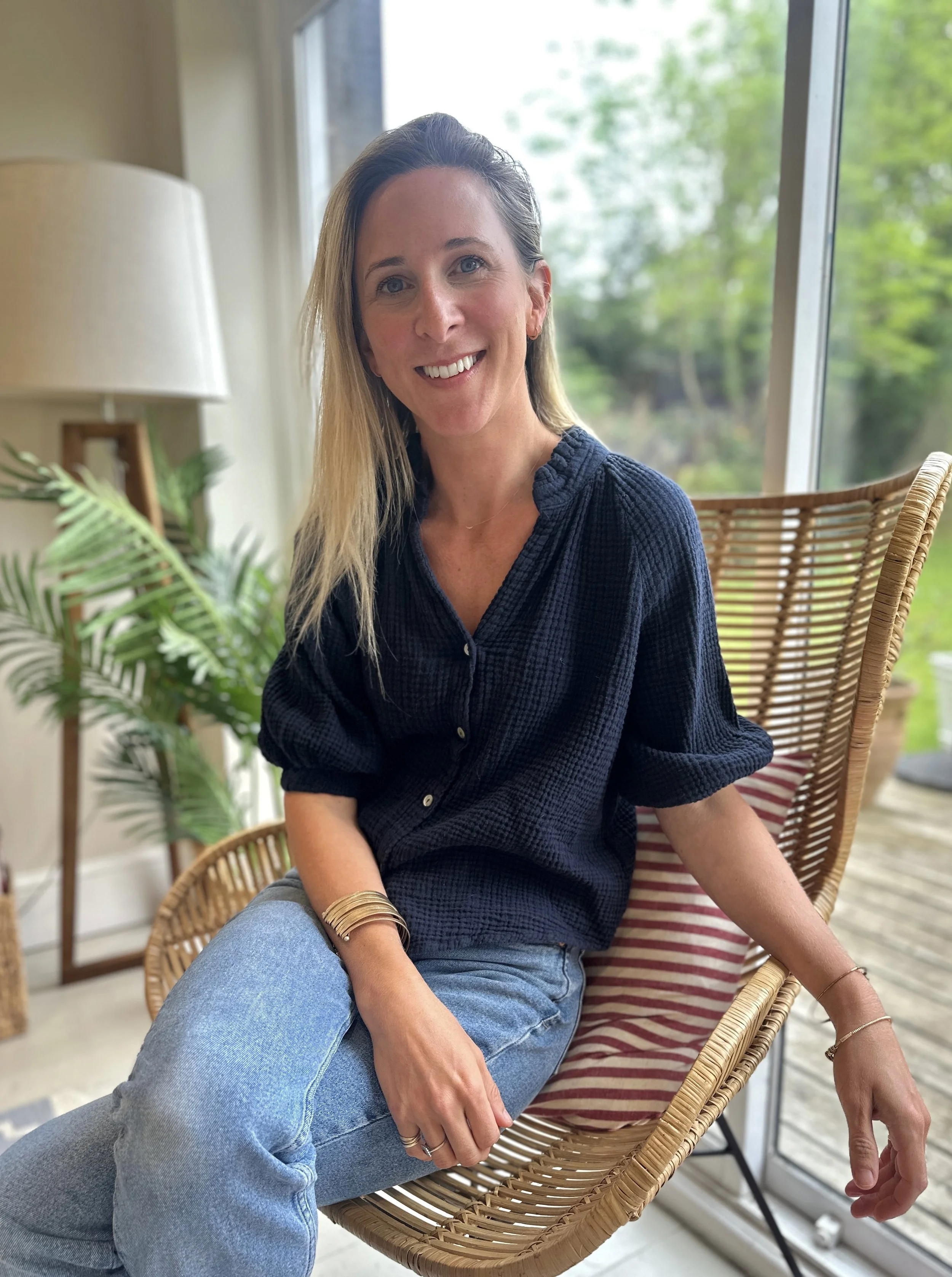 A woman with blonde hair and blue eyes sitting on a wicker chair in front of a large window with greenery outside, smiling at the camera.