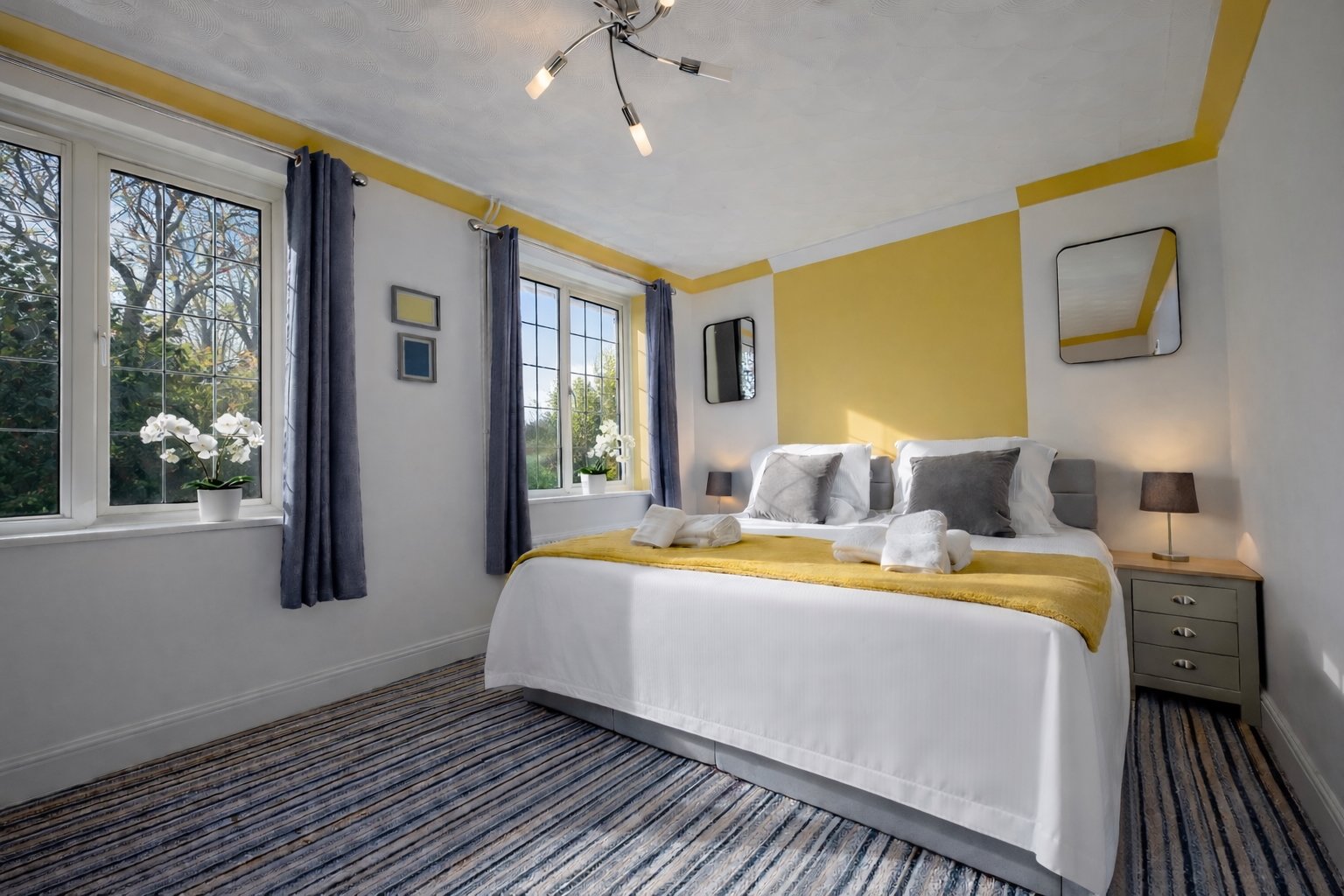 A cozy bedroom with a large bed, white linens, and gray pillows. Two windows with dark blue curtains, a yellow accent wall, and mirror decor. Nightstands on each side with black lamps. Potted orchids on window sills, striped carpet, and modern ceilin