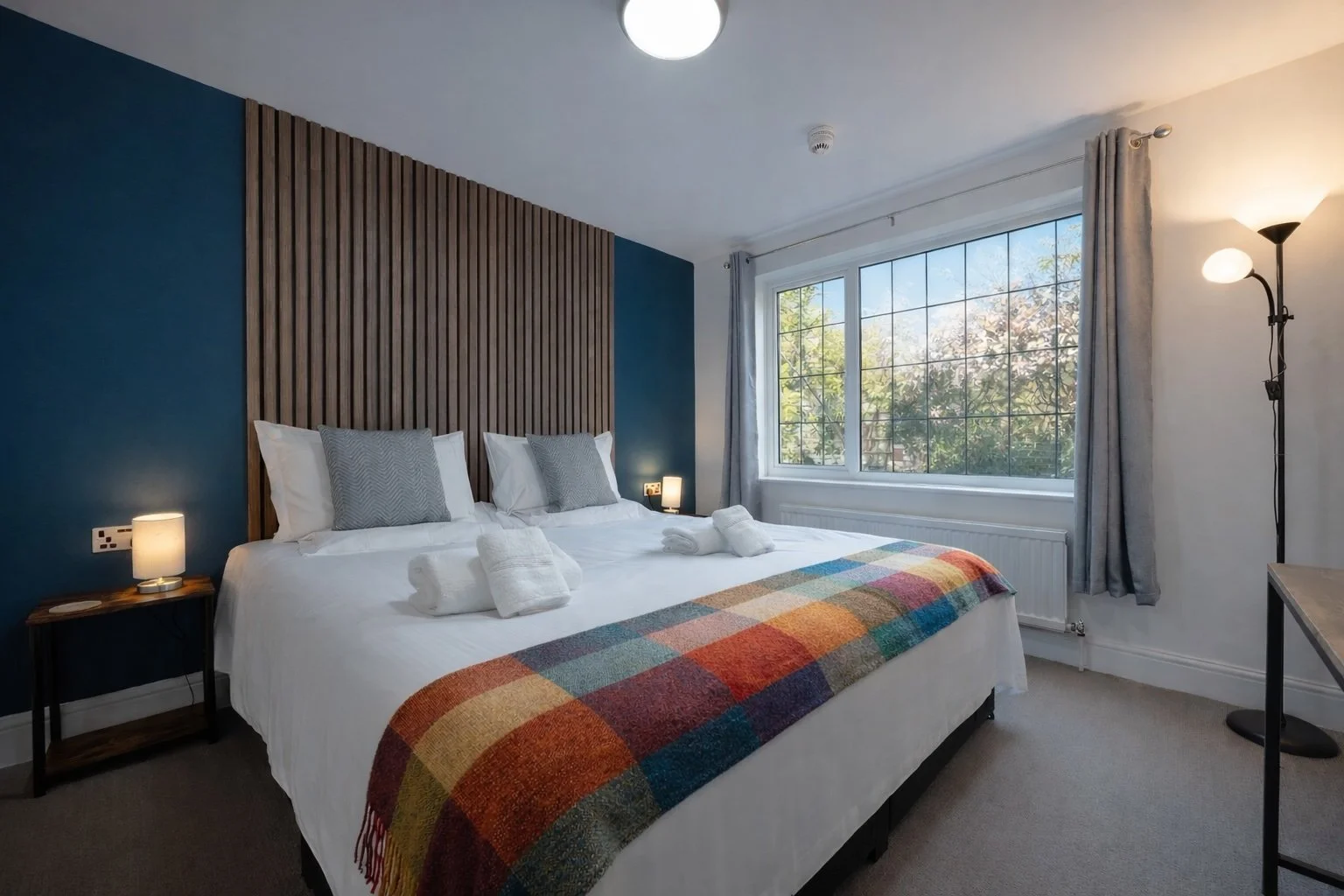 A cozy bedroom with a large bed featuring white bedding, gray pillows, and a colorful plaid throw blanket. The wall behind the bed has vertical wooden slats and is painted blue. There's a bedside table with a small lamp on each side of the bed. A lar