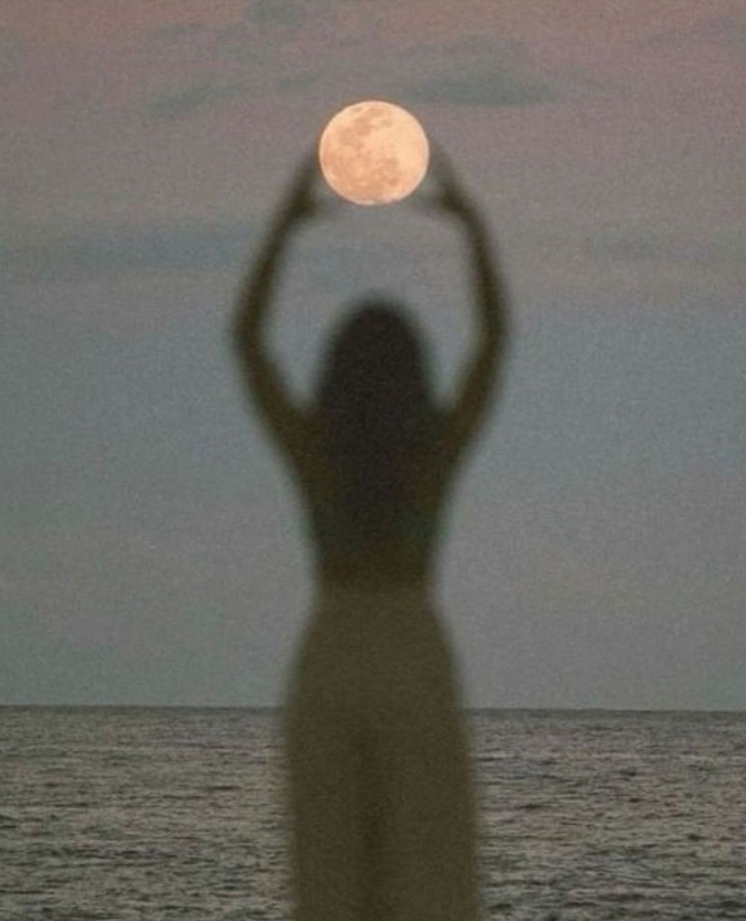 A person standing on a beach with blurred silhouette, holding their hands above their head, creating a frame for the full moon in the sky, with the ocean in the background.