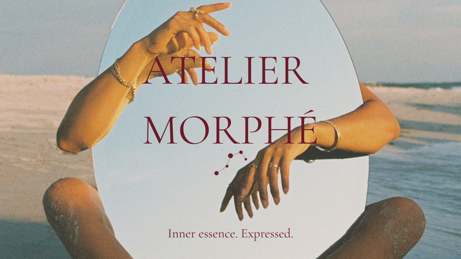 A digital image of a round mirror held by two hands, with a beach scene in the background. The mirror displays the words "ATELIER MORPHÉ" in large text and "Inner essence. Expressed." in smaller text below, with the hands adorned with rings and bracelets.