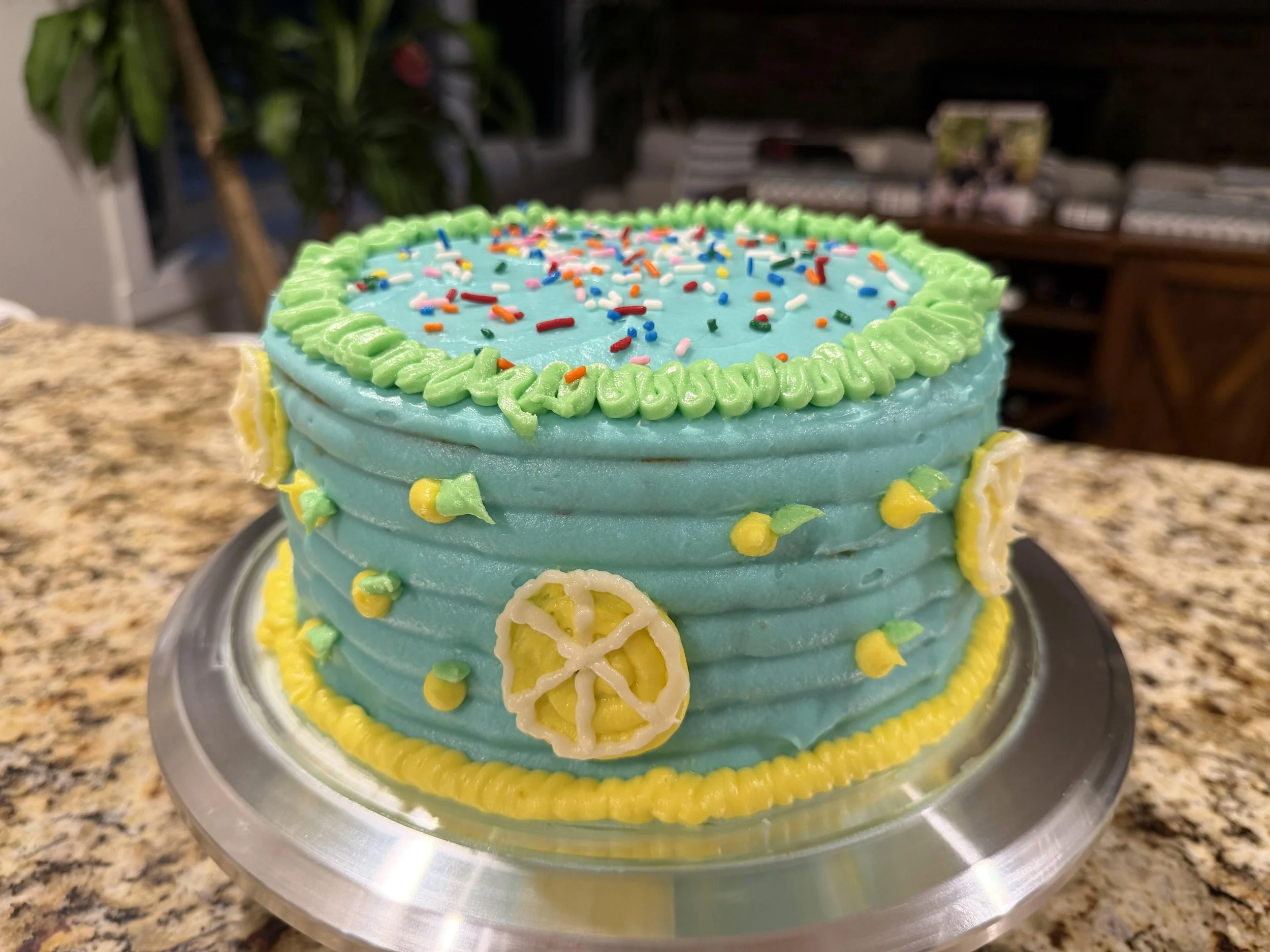 A celebratory cake with blue icing, green, yellow, white, pink, and red sprinkles, and lemon slice decorations, placed on a silver cake stand on a kitchen countertop.