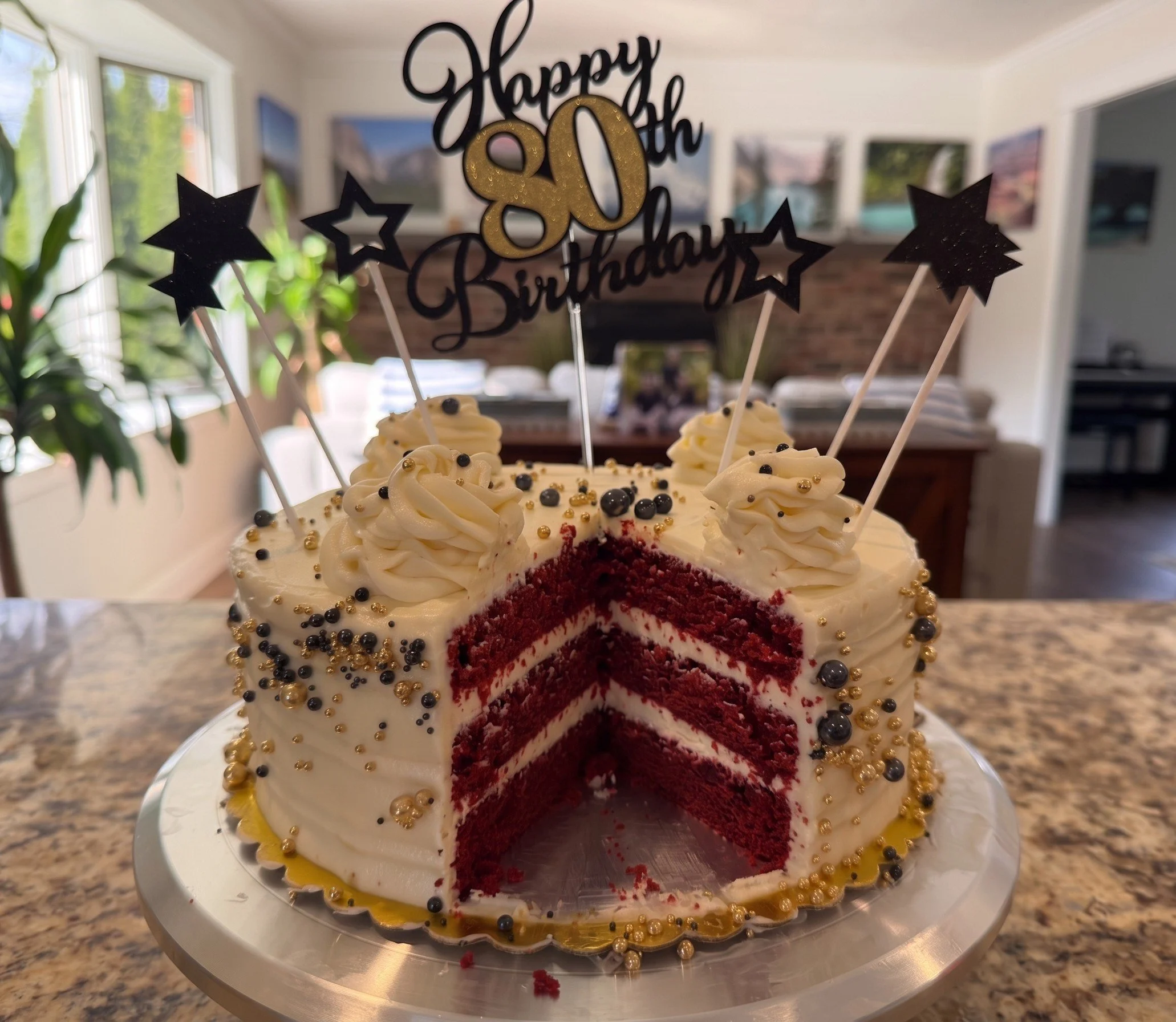 A birthday cake with white frosting and black and gold decorative sprinkles, partially sliced to reveal red velvet inside. The cake has toppers that say "Happy 80th Birthday" with stars attached to sticks, placed on a white plate on a kitchen countertop, with windows and house artwork visible in the background.