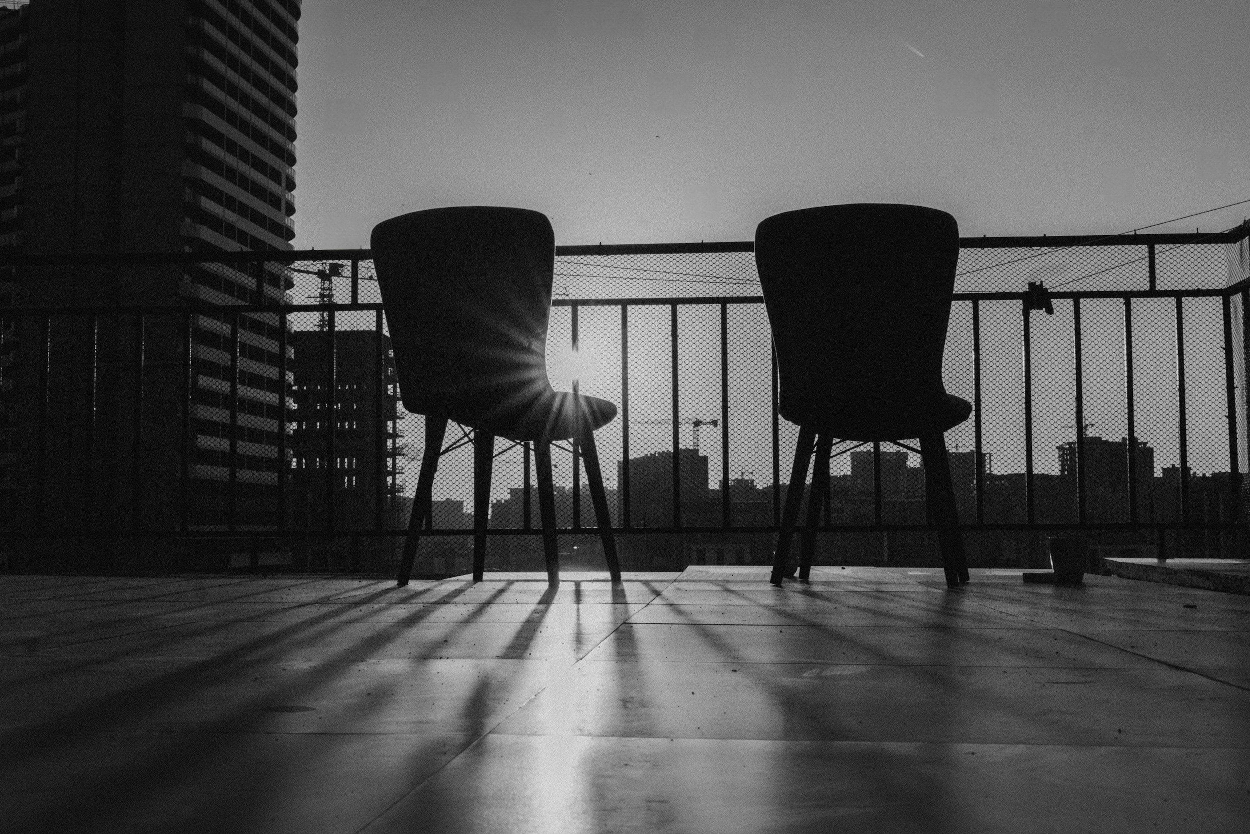 Two chairs on a rooftop or balcony with a cityscape in the background, silhouetted by the setting or rising sun.