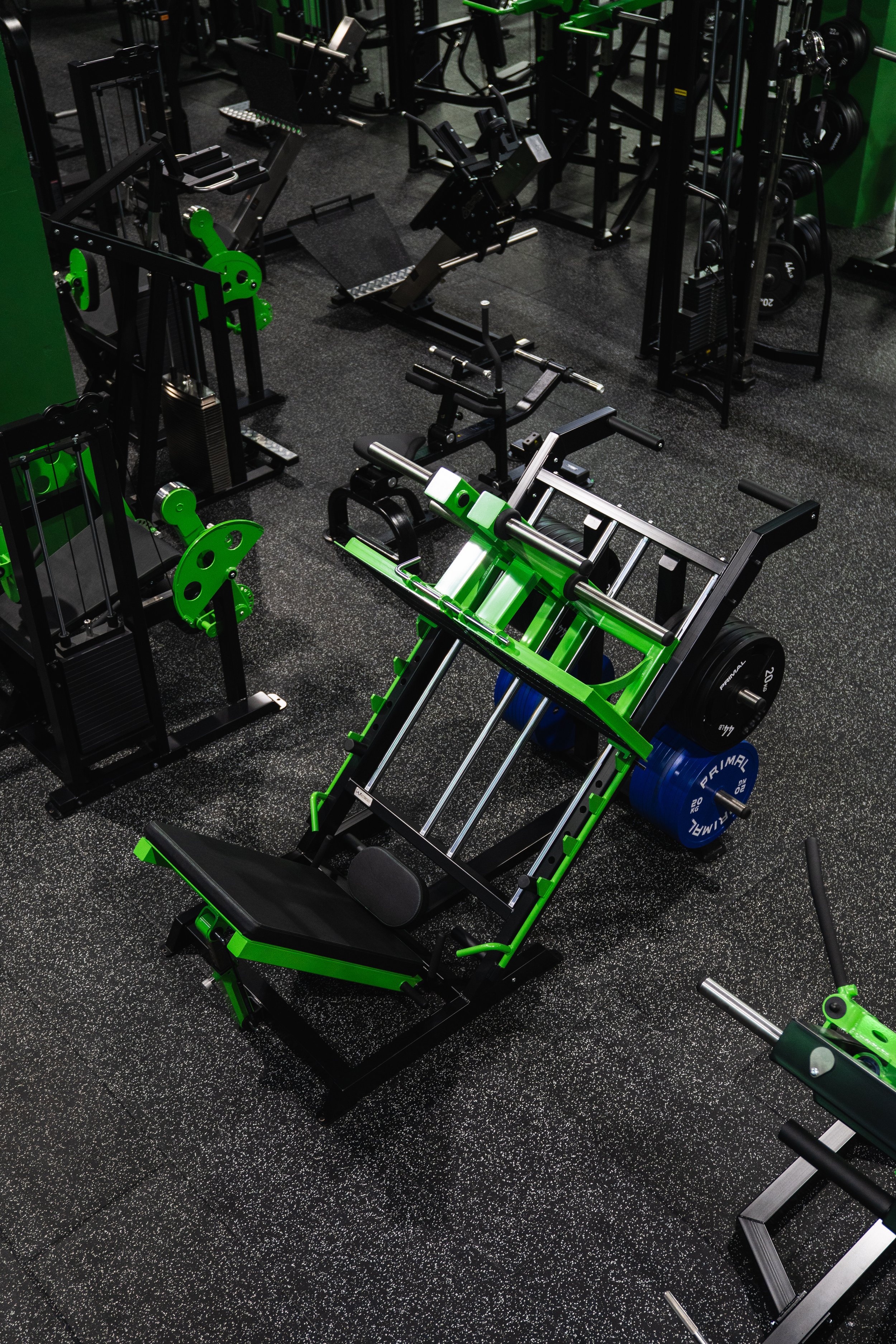 Gym equipment including weightlifting benches, barbells, weight plates, and resistance machines in a gym with black and green color accents.
