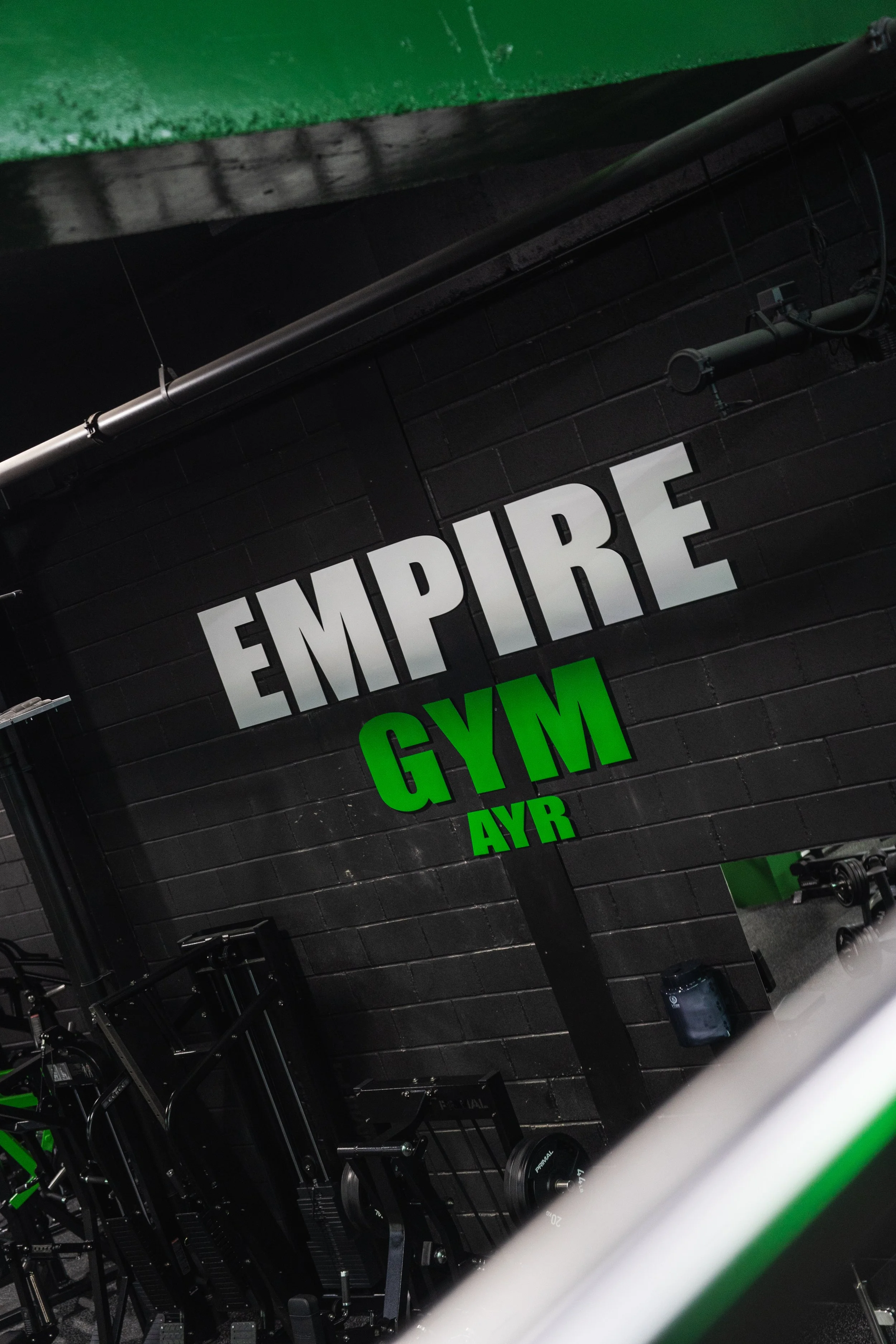Gym interior with black brick wall, white and green sign reading 'EMPire GYM'. Fitness equipment, weights, and a black clean floor.