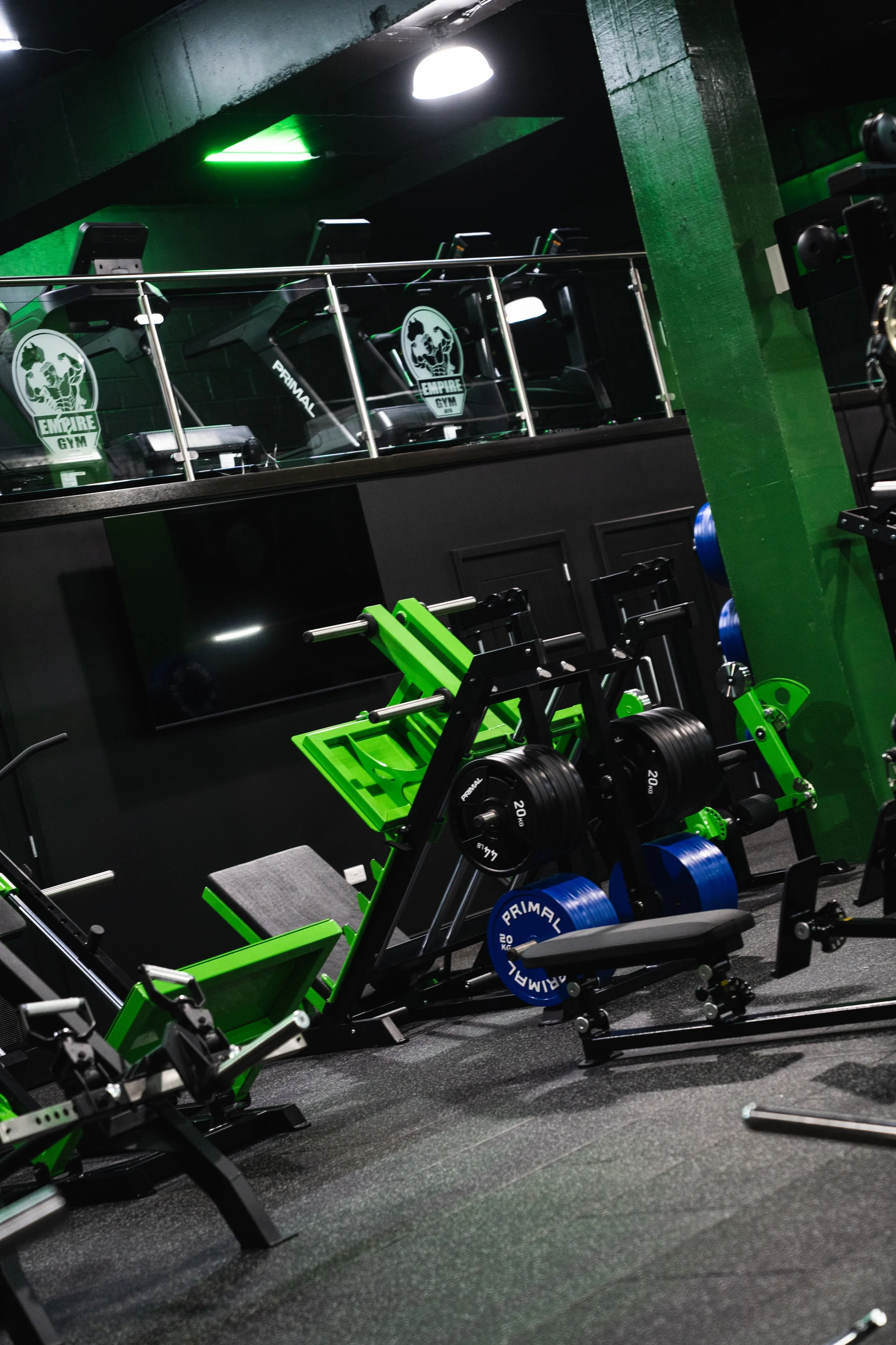 Empty gym with various workout equipment including dumbbells, squat racks, and cardio machines. The gym has black flooring and green accents, with a glass balcony overlooking the workout area and a green and black color scheme.