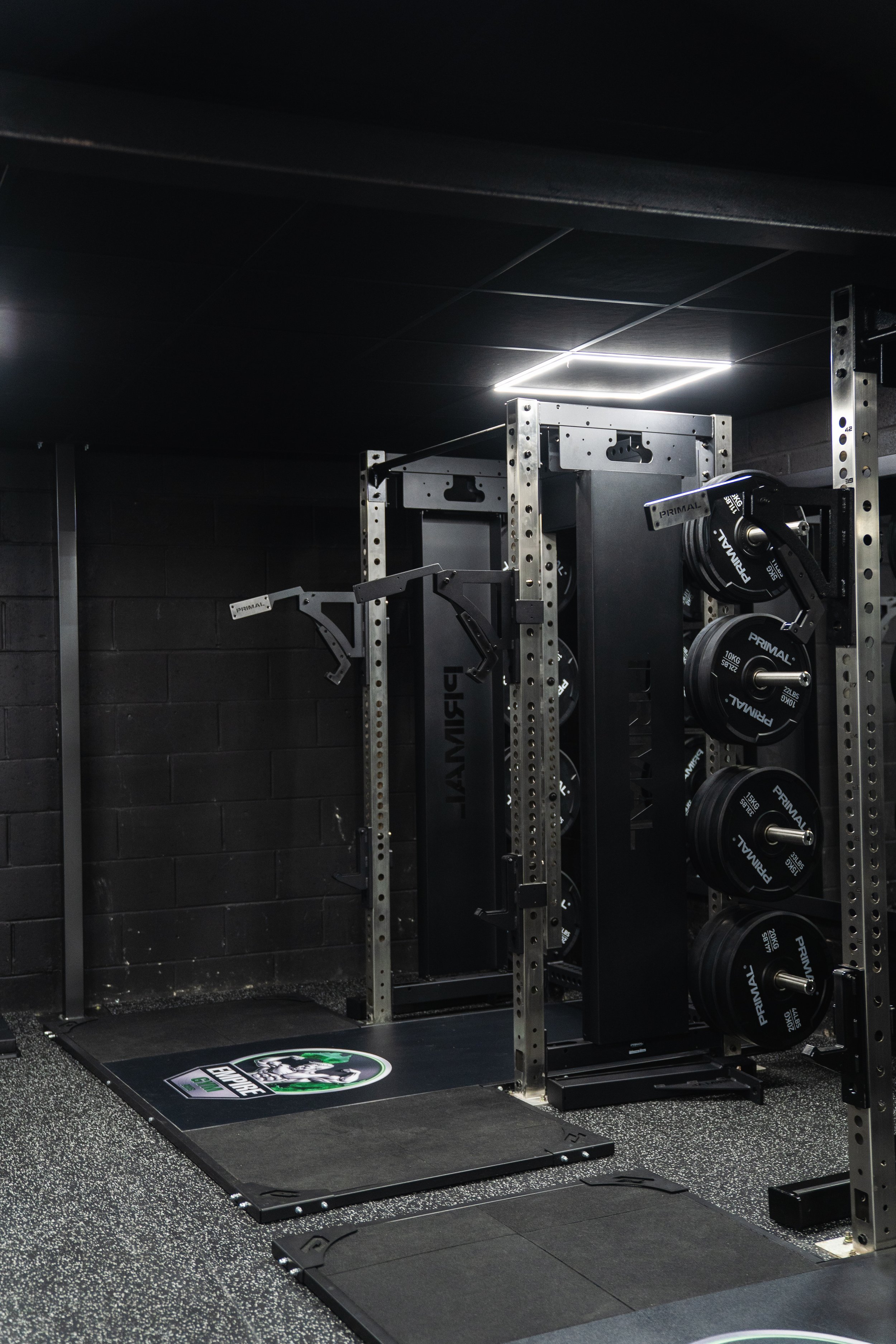 A gym setup with a squat rack, weight plates, and a lifting platform on a speckled black and gray floor. The room has dark walls and ceiling.