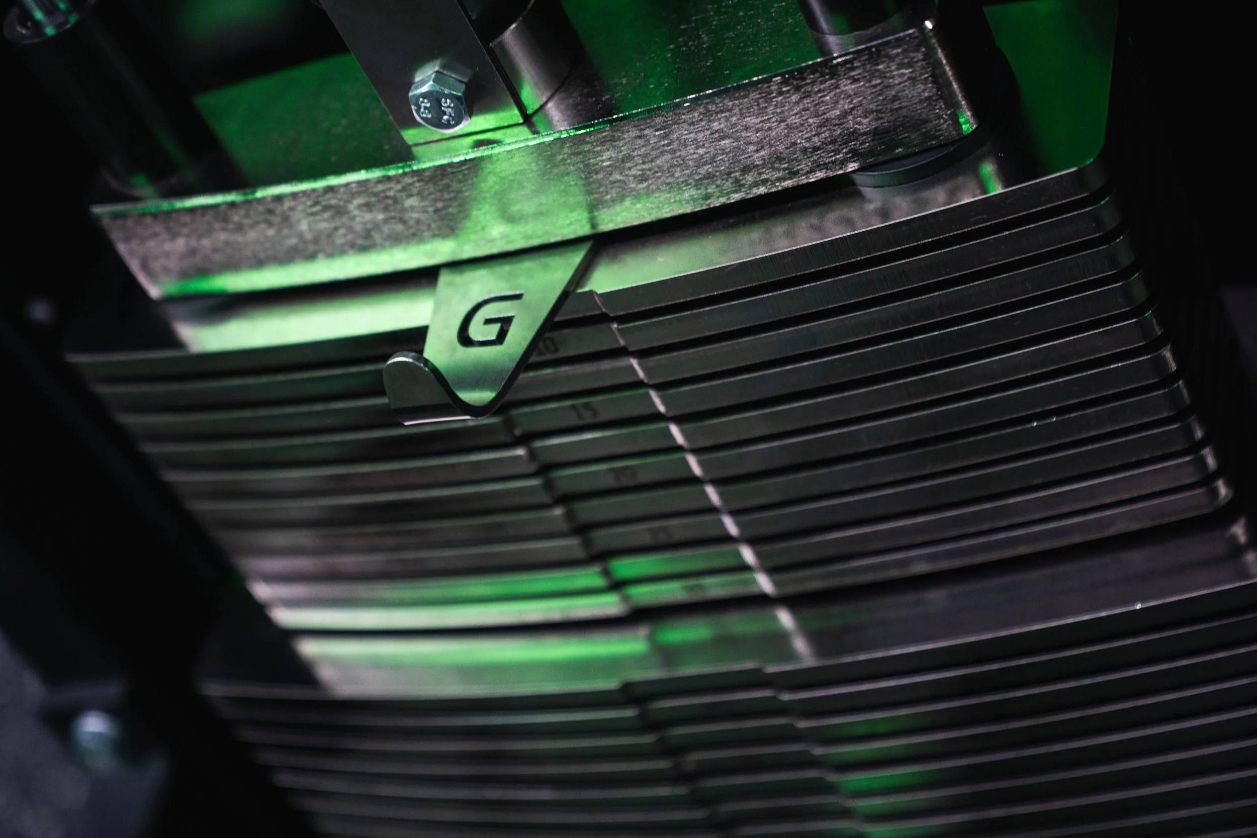 Close-up of a computer processor with a metal heat sink and a small metallic clip marked with the letter 'G' securing the heatsink to the CPU, with green light reflections highlighting the shiny metallic surfaces.