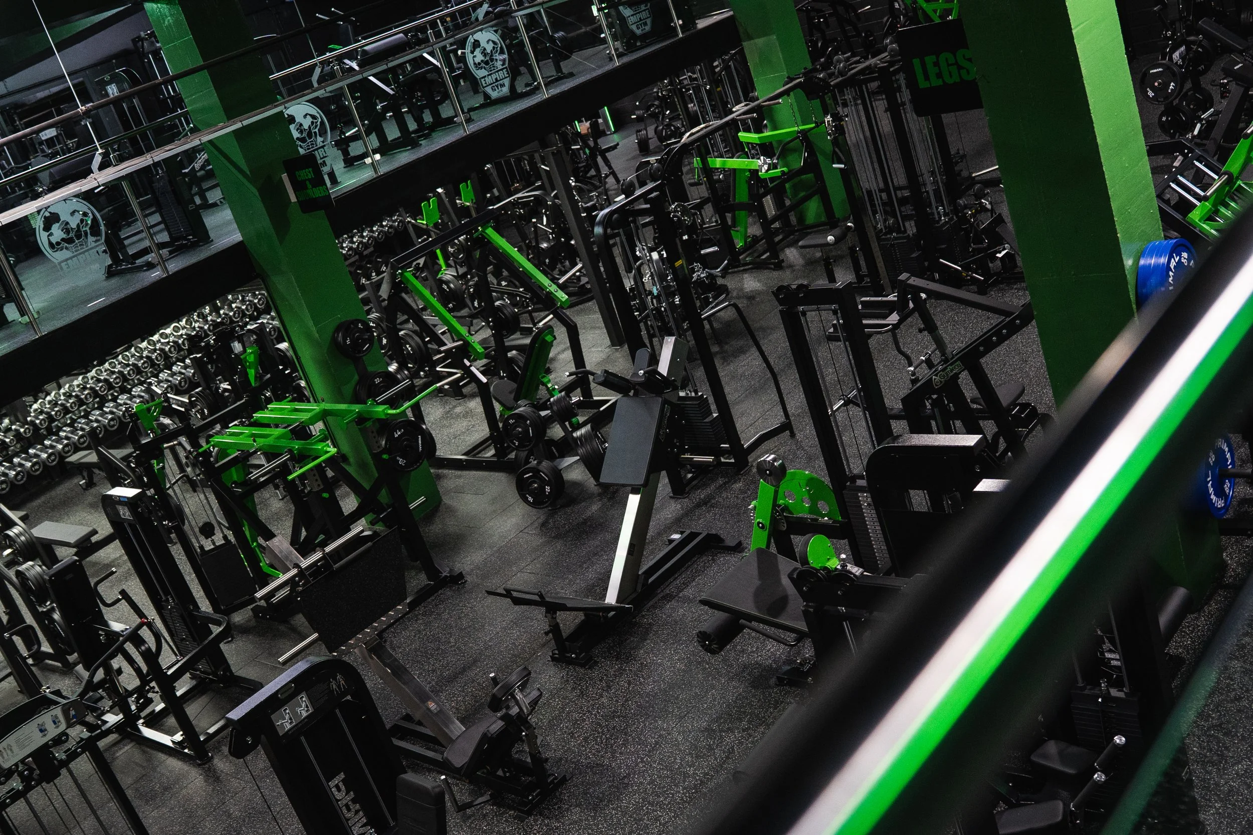 Empty gym with various workout equipment, including dumbbells, weight machines, and benches, with green accents and a balcony area.
