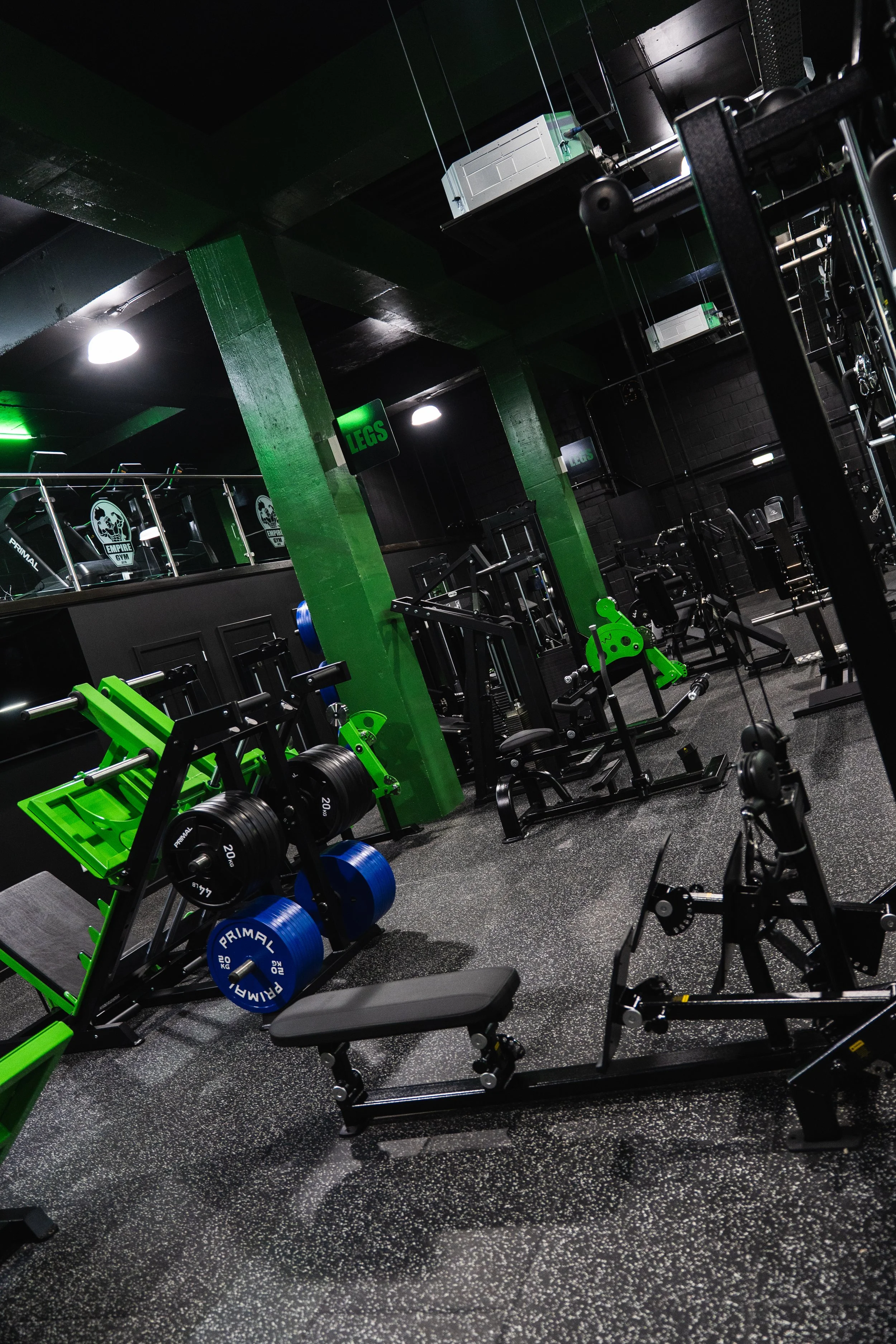 Inside a gym with various strength training equipment, including weight racks, benches, and machines, with green accents and black walls.