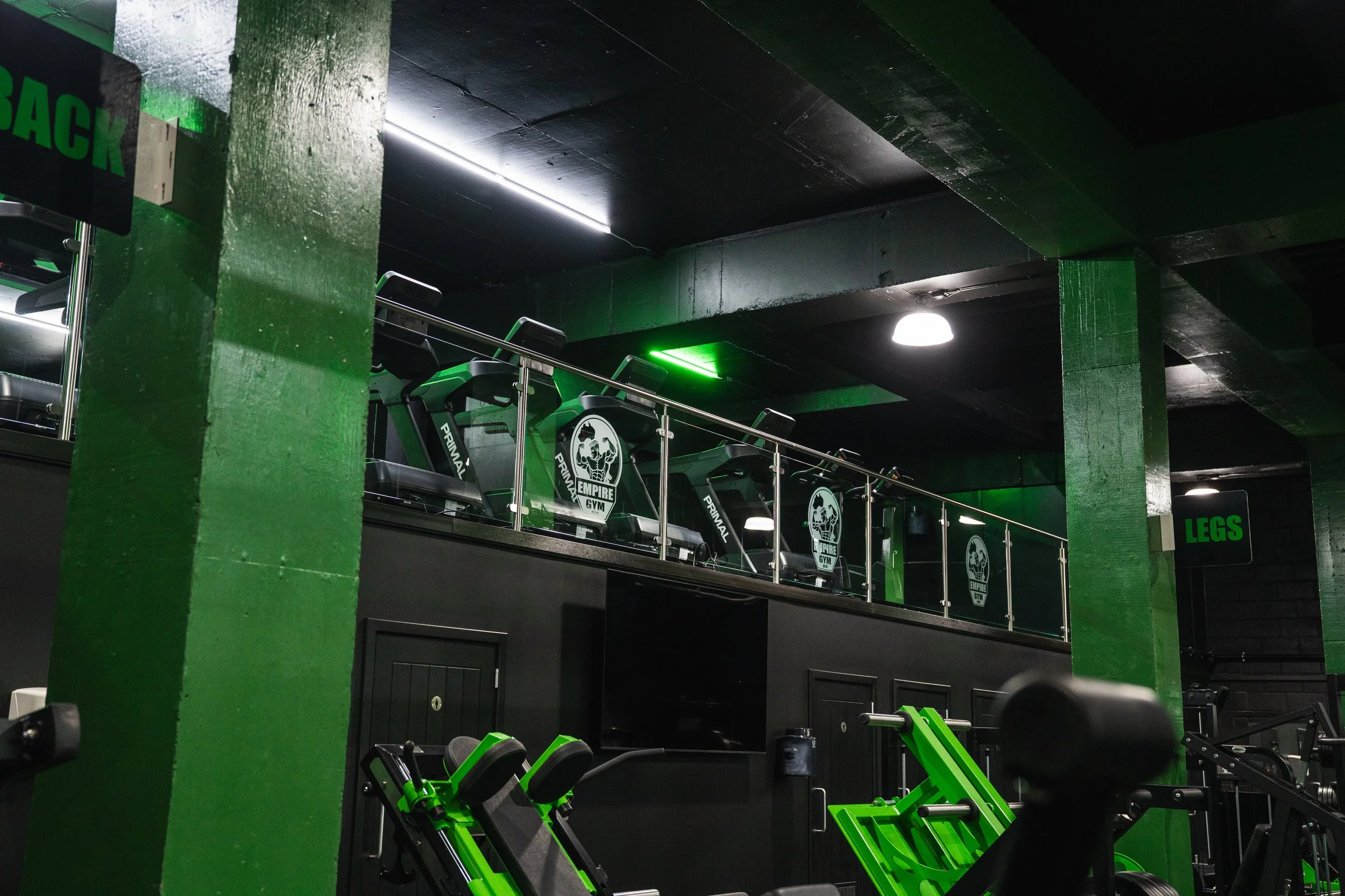 Interior of a gym with green and black color scheme. Equipment includes treadmill machines with Empire Gym logos, a staircase with green railings, and gym sections labeled 'BACK' and 'LEGS'. Green lighting accentuates the space.