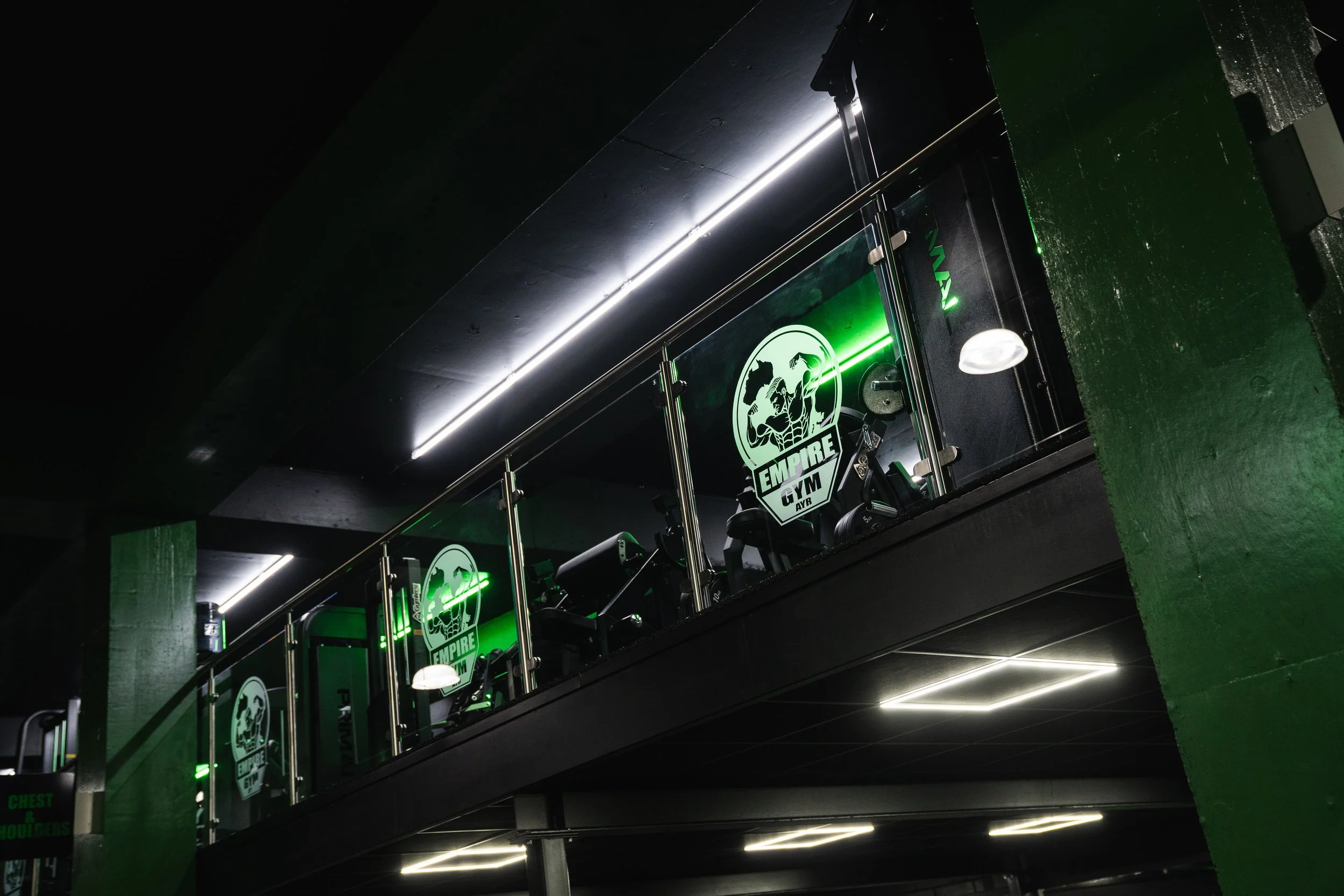 Interior of a gym with black walls and ceiling, green accents, and branding for Empire Gym. There is a mirrored wall with workout equipment reflected, and neon green lights highlight the space.