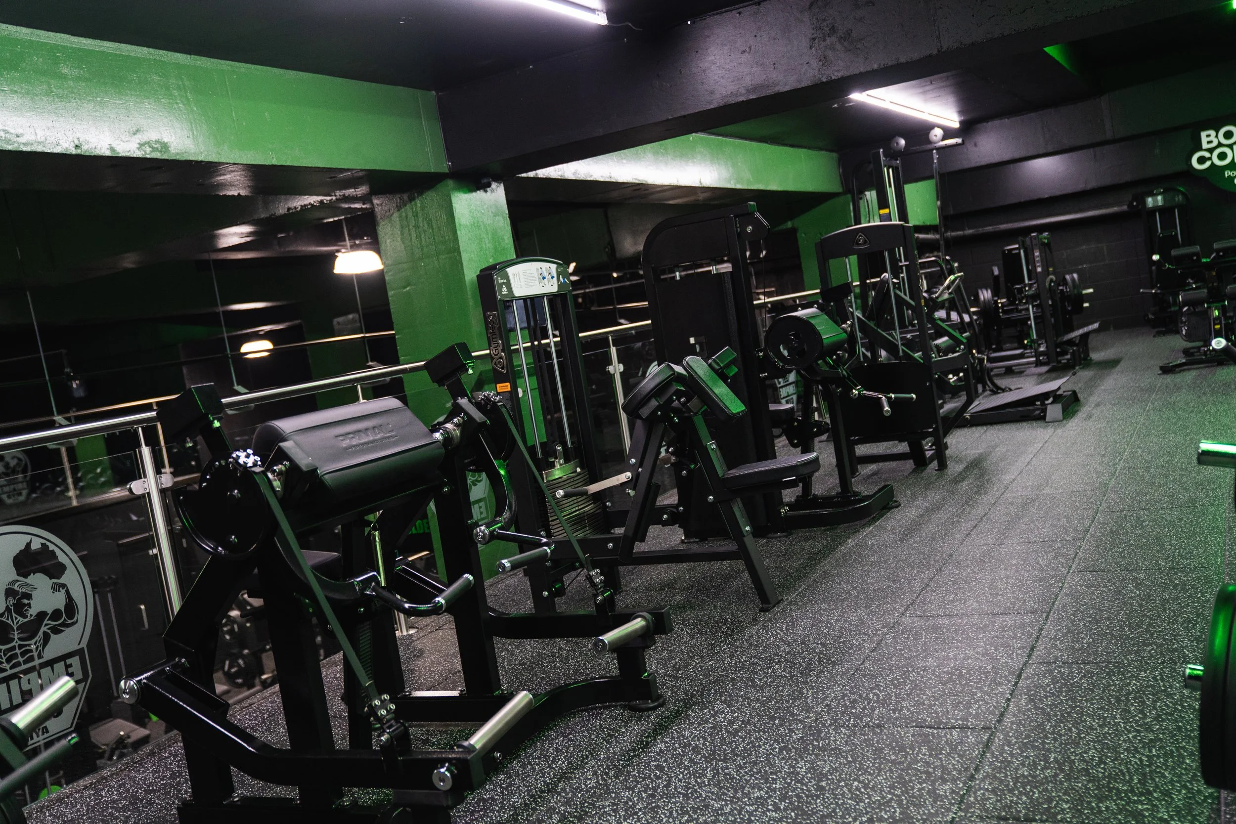 A gym with various workout machines, including seated leg press and elliptical trainers, in a room with black and green walls and a speckled black floor, illuminated by fluorescent lights.