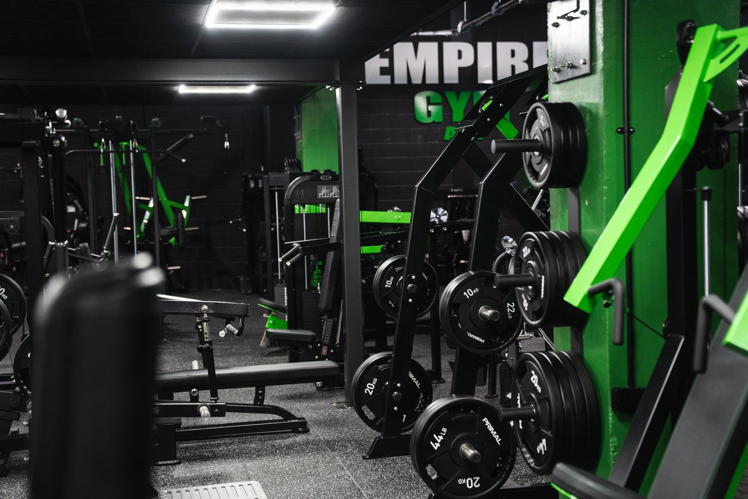 Interior of a gym with various workout equipment, including weights and machines, with a black and green color scheme and a sign that reads 'Empire Gym' on the wall.